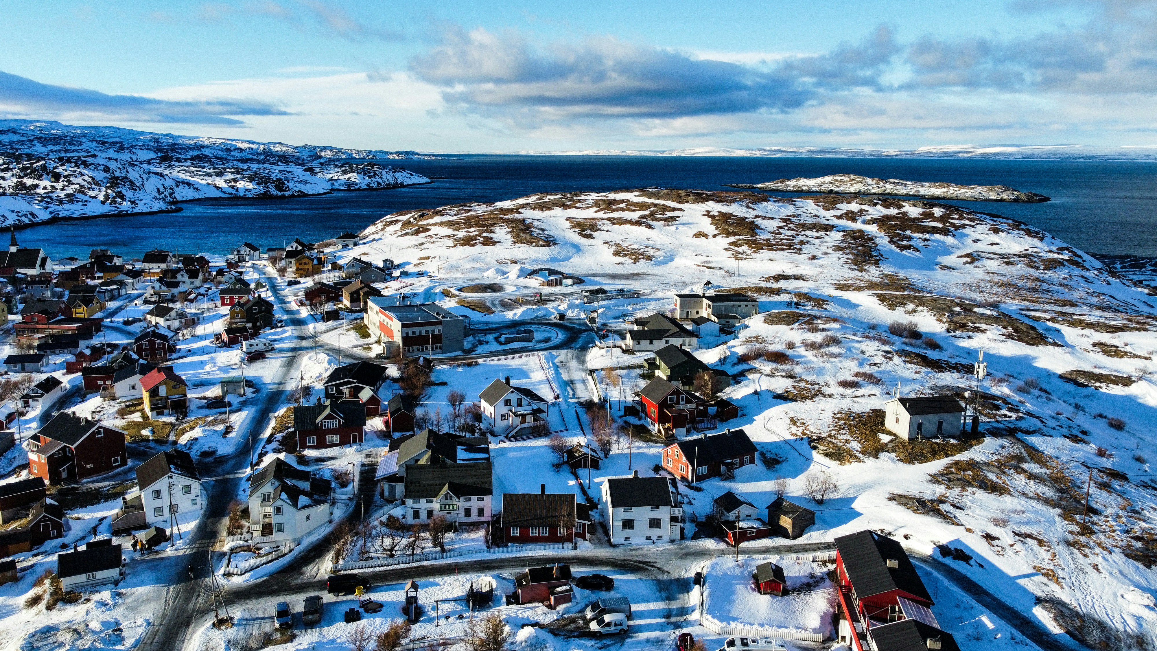 An aerial view of a small town covered in snow during winter, houses are scattered, the streets are also covered with thick snow, and there is a blue sea in the distance. - free wallpaper image