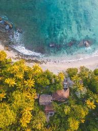 An aerial view of a house surrounded by green trees on the coast, with a sandy beach and clear blue water nearby. - free wallpaper image