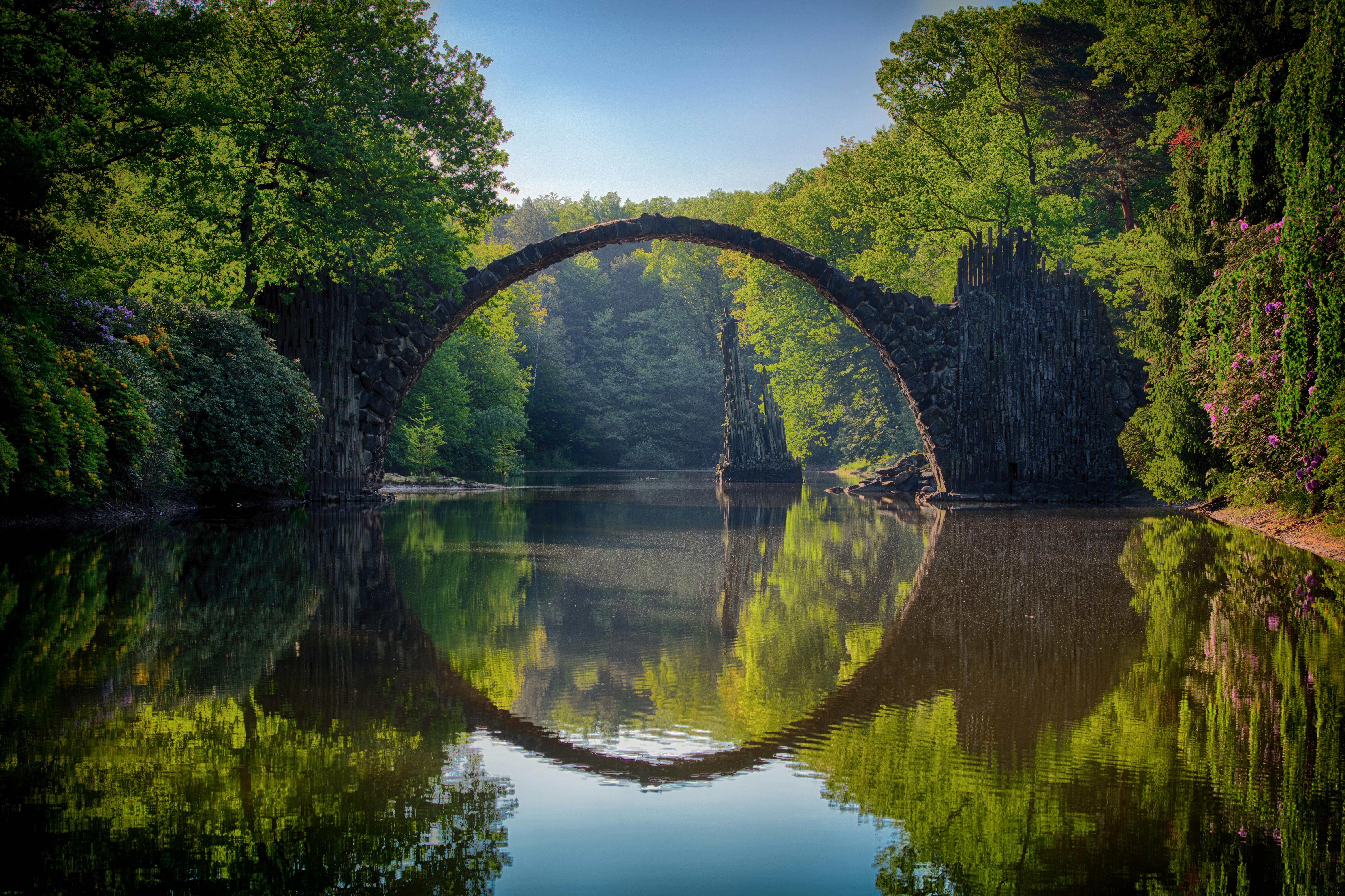 An ancient stone arch bridge spans a tranquil lake, with lush trees lining the banks, the reflections are clearly visible, the scene is peaceful and serene. - free wallpaper image