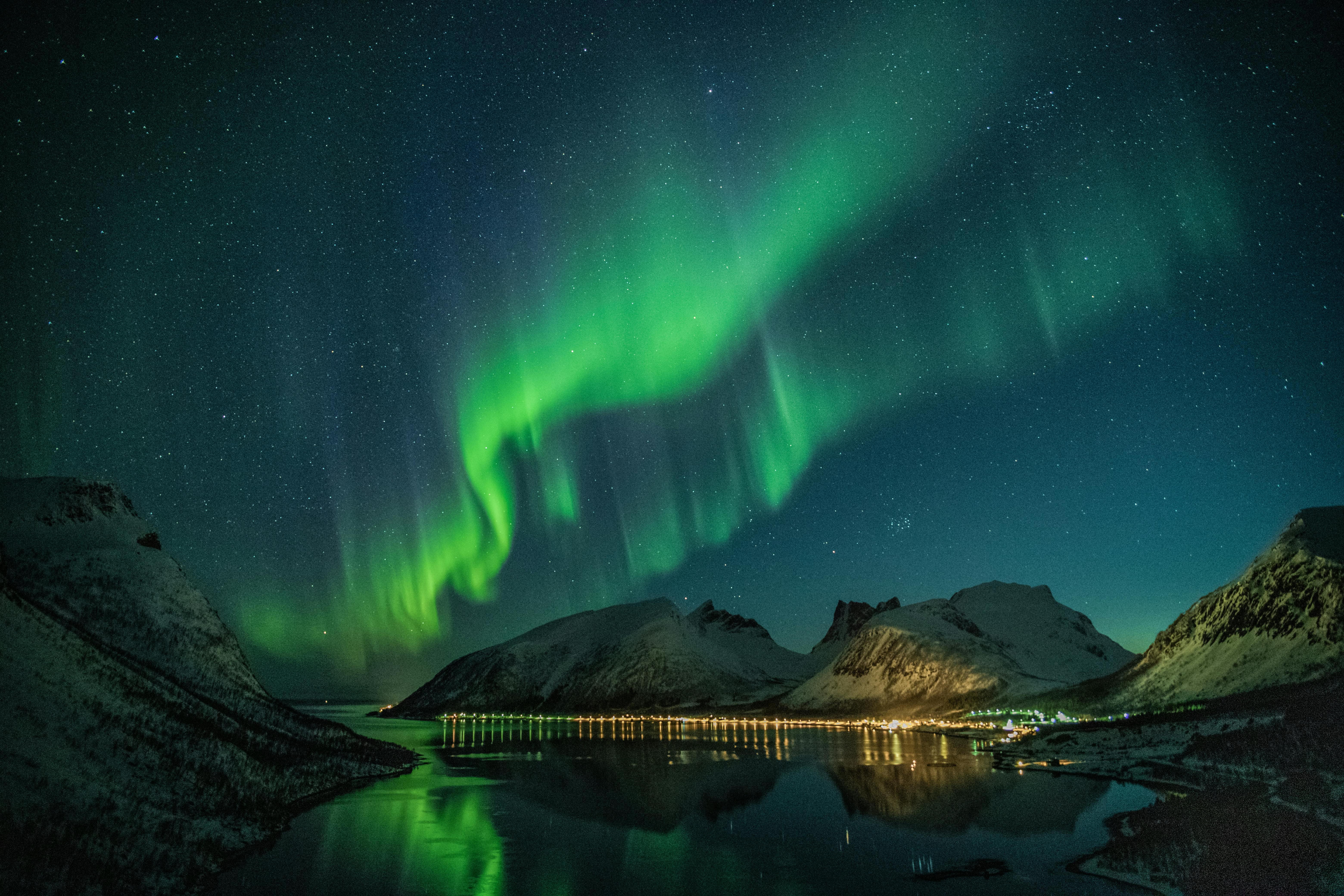 The aurora borealis is visible in the night sky, illuminating the mountains and lake with green light. There are also scattered lights in the distance. - free wallpaper image