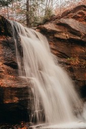 A waterfall cascades down a rocky cliff, the water rushing over the edge. Surrounding trees and rocks are displaying autumnal colors. - free wallpaper image