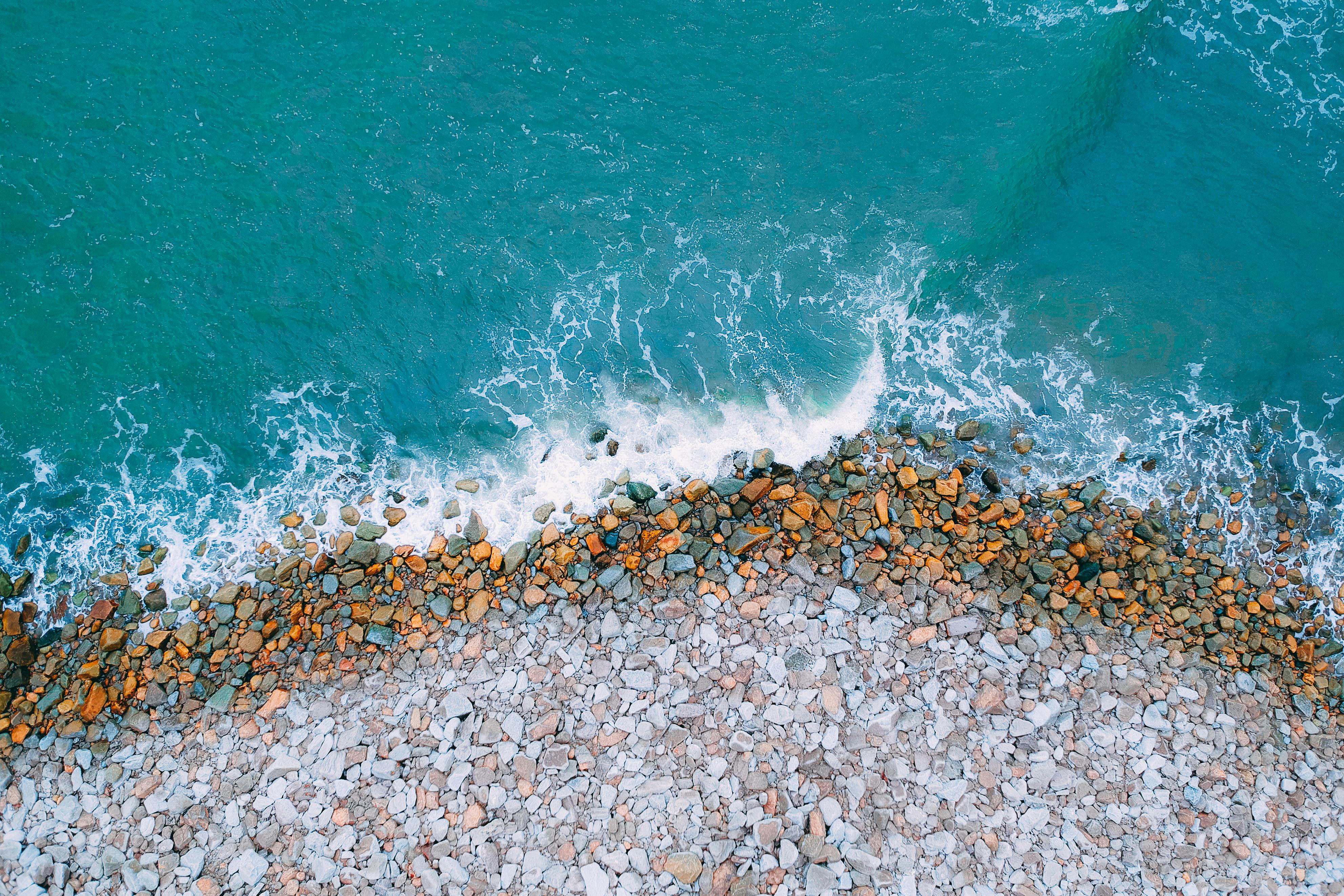 An aerial view of the coastline, where waves crash against the shore, white foam contrasting with blue water, the coast is covered with pebbles of different sizes. - free wallpaper image