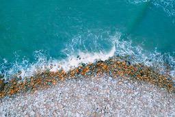 An aerial view of the coastline, where waves crash against the shore, white foam contrasting with blue water, the coast is covered with pebbles of different sizes. - free wallpaper image