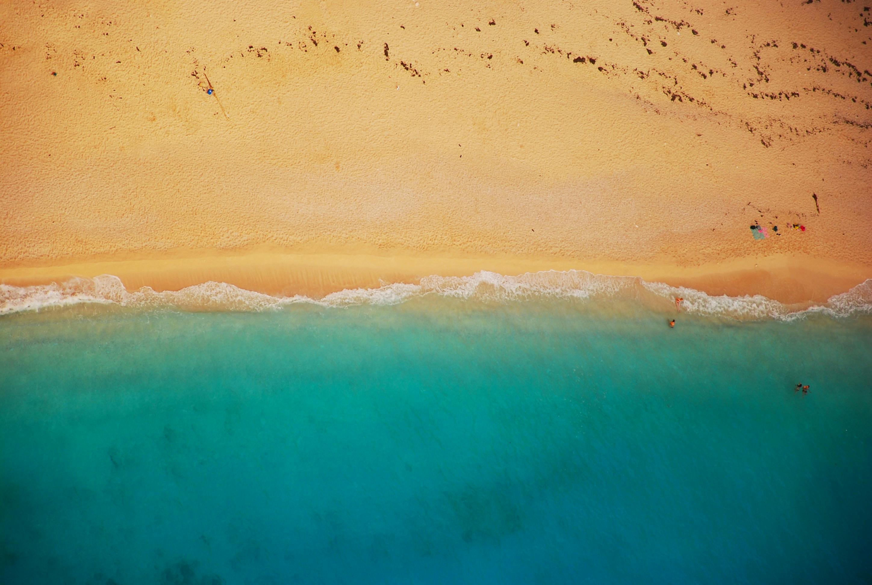 An aerial view of a sandy beach meeting the turquoise blue ocean, with a few people enjoying the water. - free wallpaper image