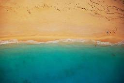 An aerial view of a sandy beach meeting the turquoise blue ocean, with a few people enjoying the water. - free wallpaper image