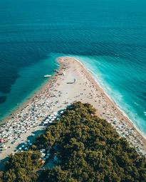 An overhead view of a beach with a full view of the beach, full of people and white umbrellas, clear blue water and surrounded by green vegetation. - free wallpaper image