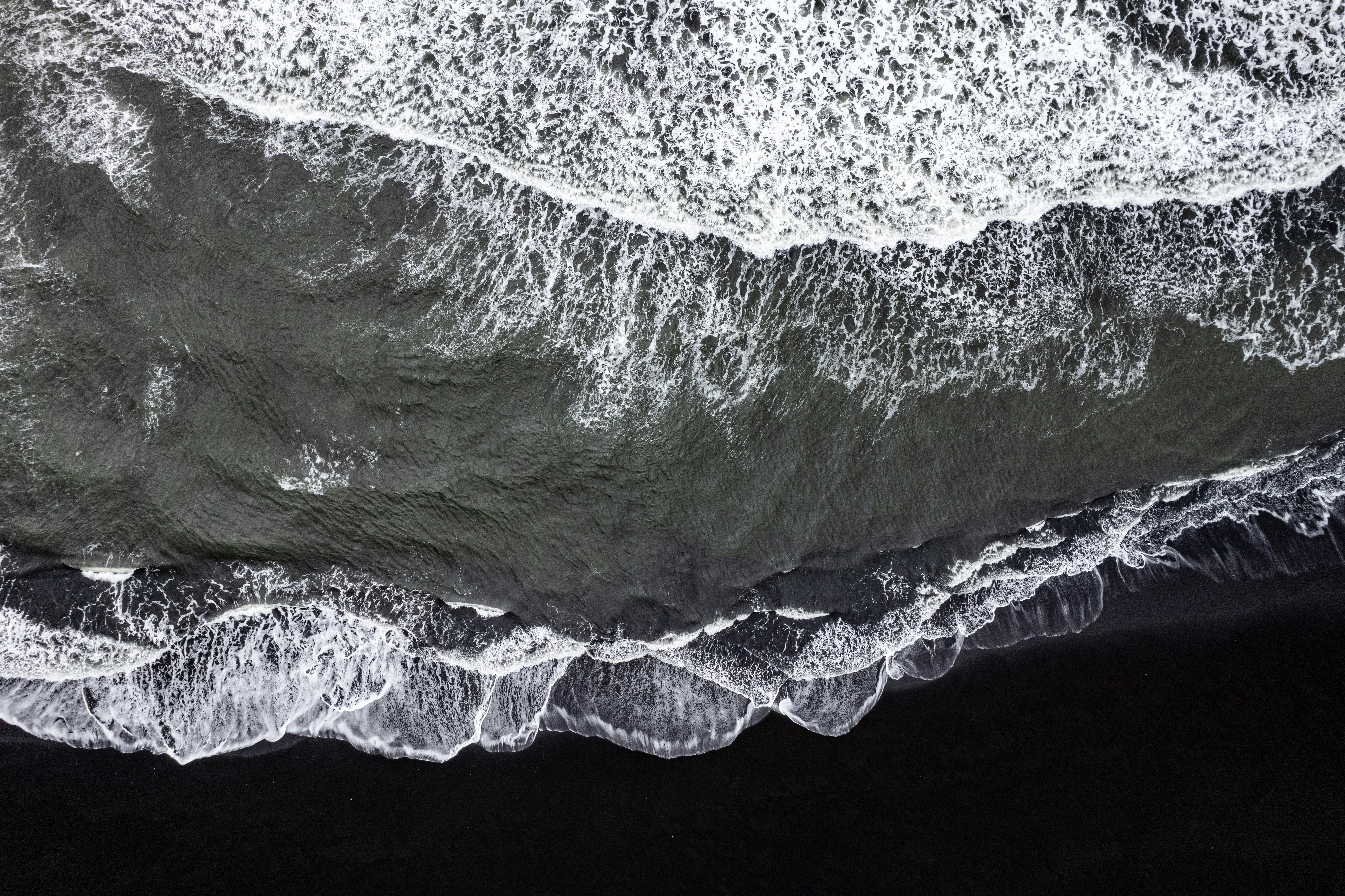 An aerial view of a black sand beach with white waves crashing against the shore. The water is a deep gray color. - free wallpaper image