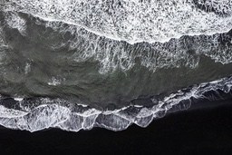 An aerial view of a black sand beach with white waves crashing against the shore. The water is a deep gray color. - free wallpaper image