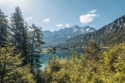 On a sunny day, a clear blue lake is surrounded by dense forests, with snow-capped mountains in the distance. - free wallpaper image