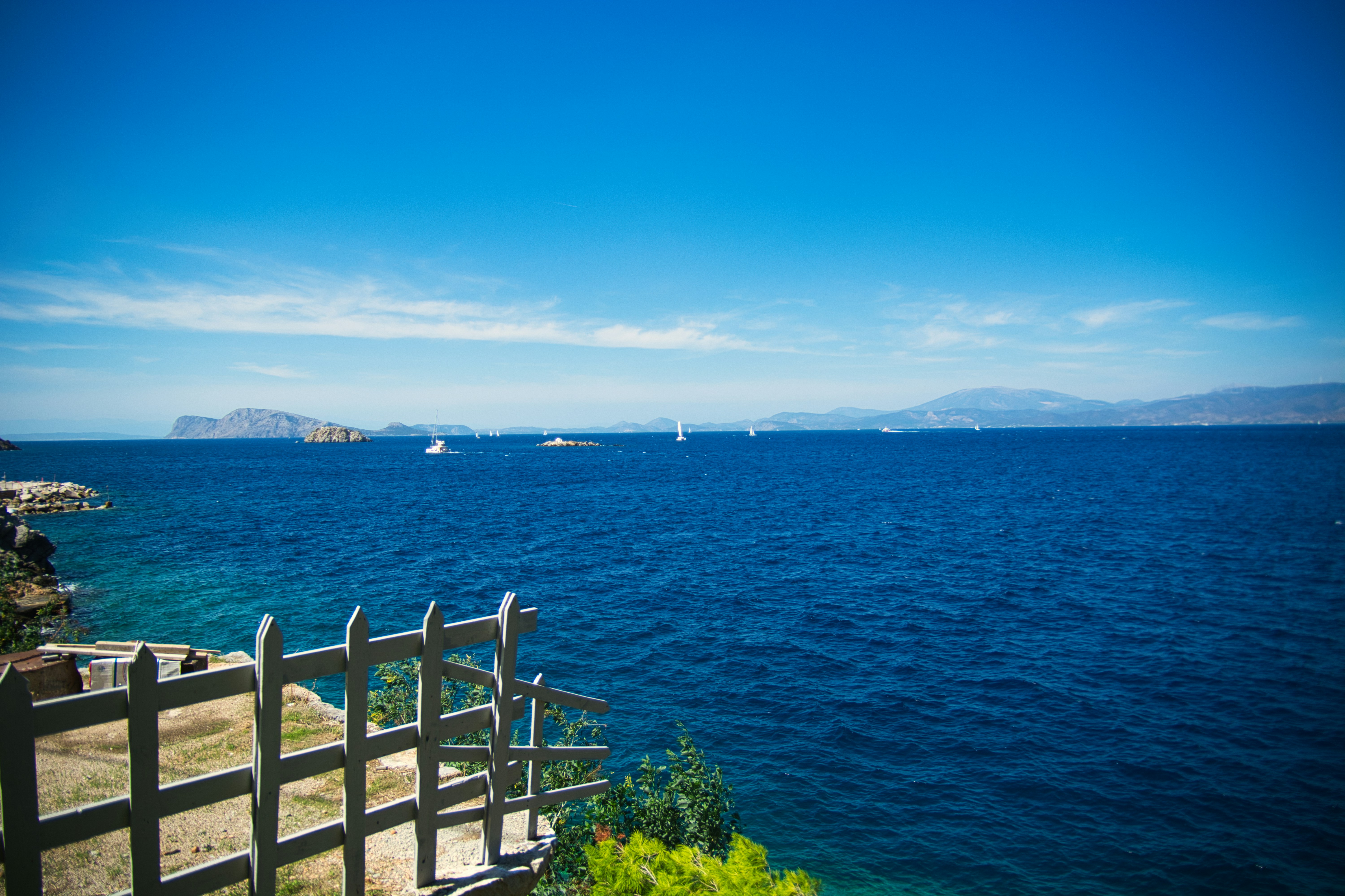 A view of the sea with blue water and white fence in front of green bushes on the shore. - free wallpaper image