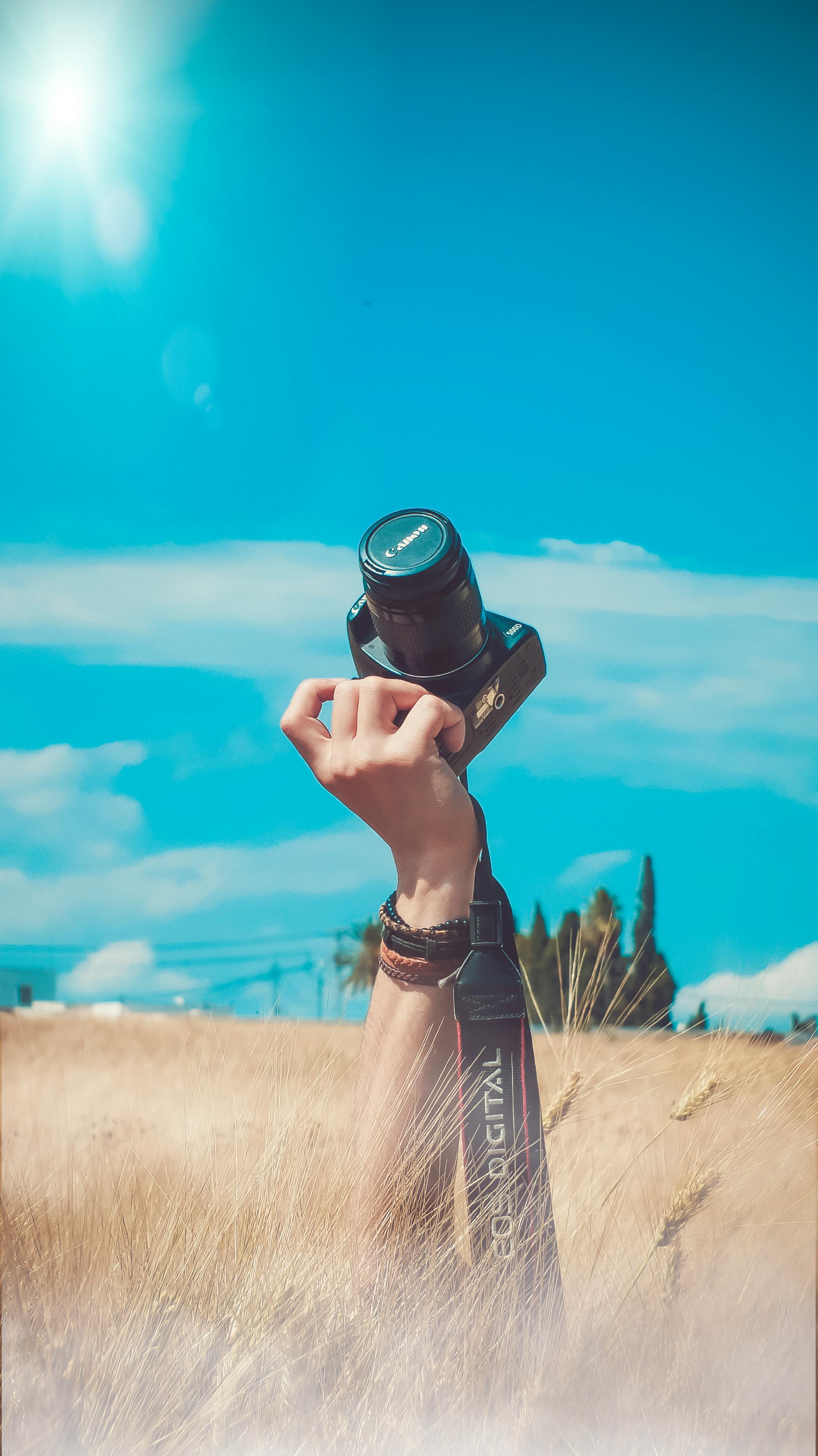 A hand holds a camera high in the air, against a backdrop of blue sky, white clouds and golden wheat fields. The camera is pointing at the sky, as if taking a picture of the beautiful sky. - free wallpaper image