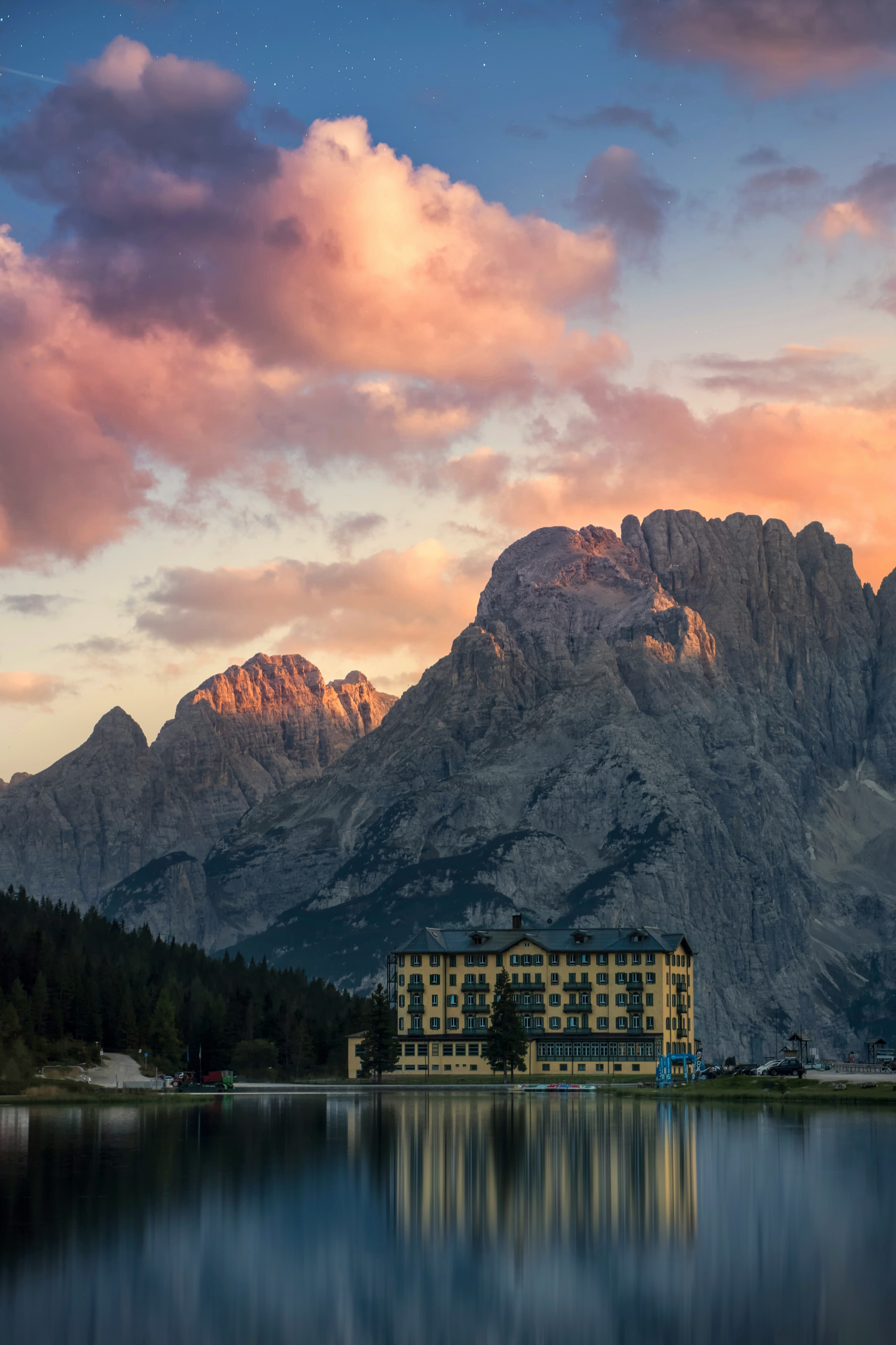 A building stands by a mountain lake, the afterglow of the sunset shines on the clouds and mountains, the lake is calm and reflects the shadows of the building and mountains. - free wallpaper image