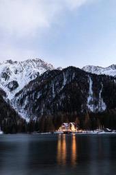 A cabin with warm lights is surrounded by snow-capped mountains in a lake, the lake surface is calm and reflects the silhouettes of mountains and houses. - free wallpaper image