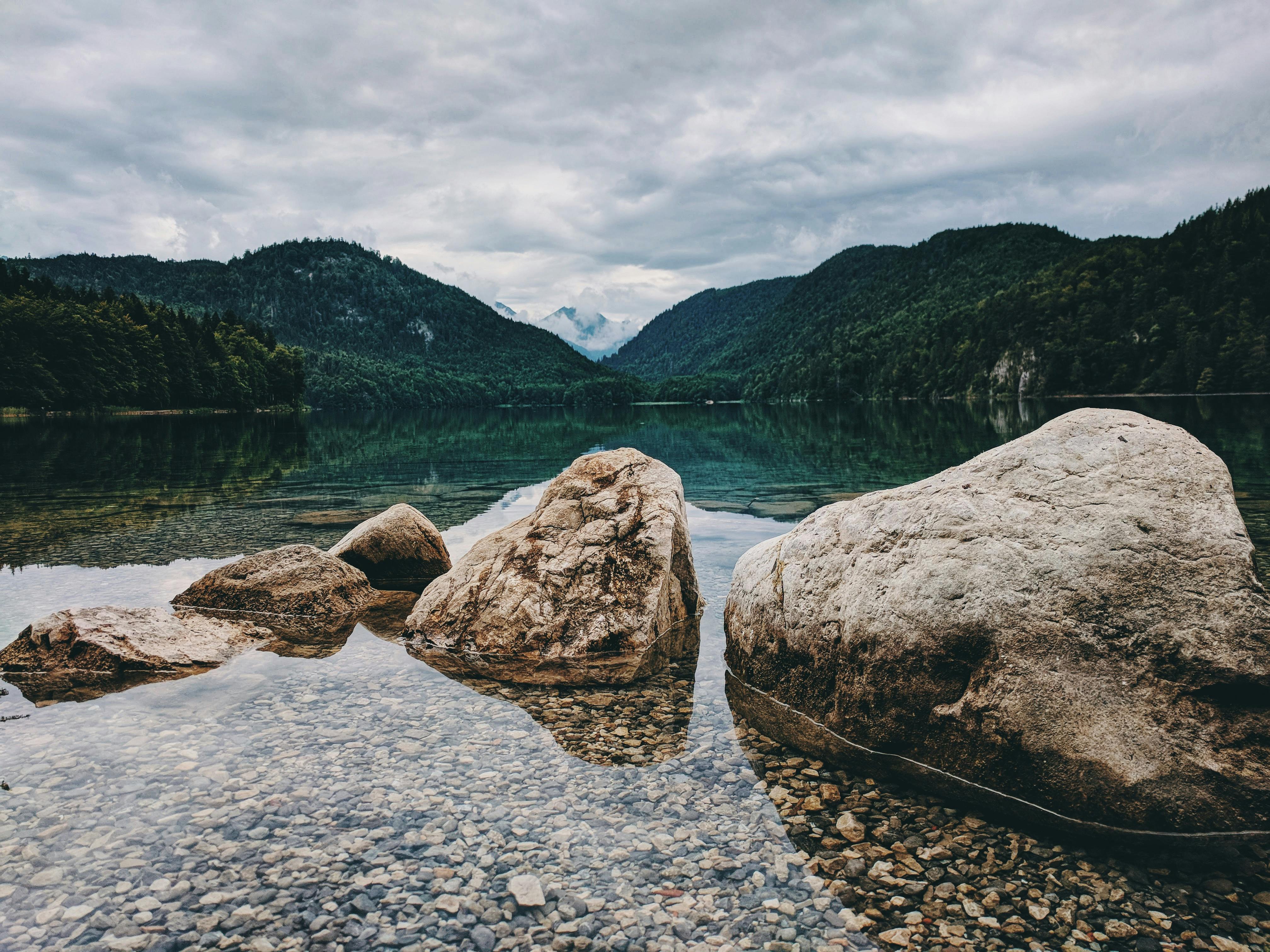 A calm lake with a few rocks scattered in the water, the mountains in the distance are reflected in the lake, the sky is full of clouds. - free wallpaper image