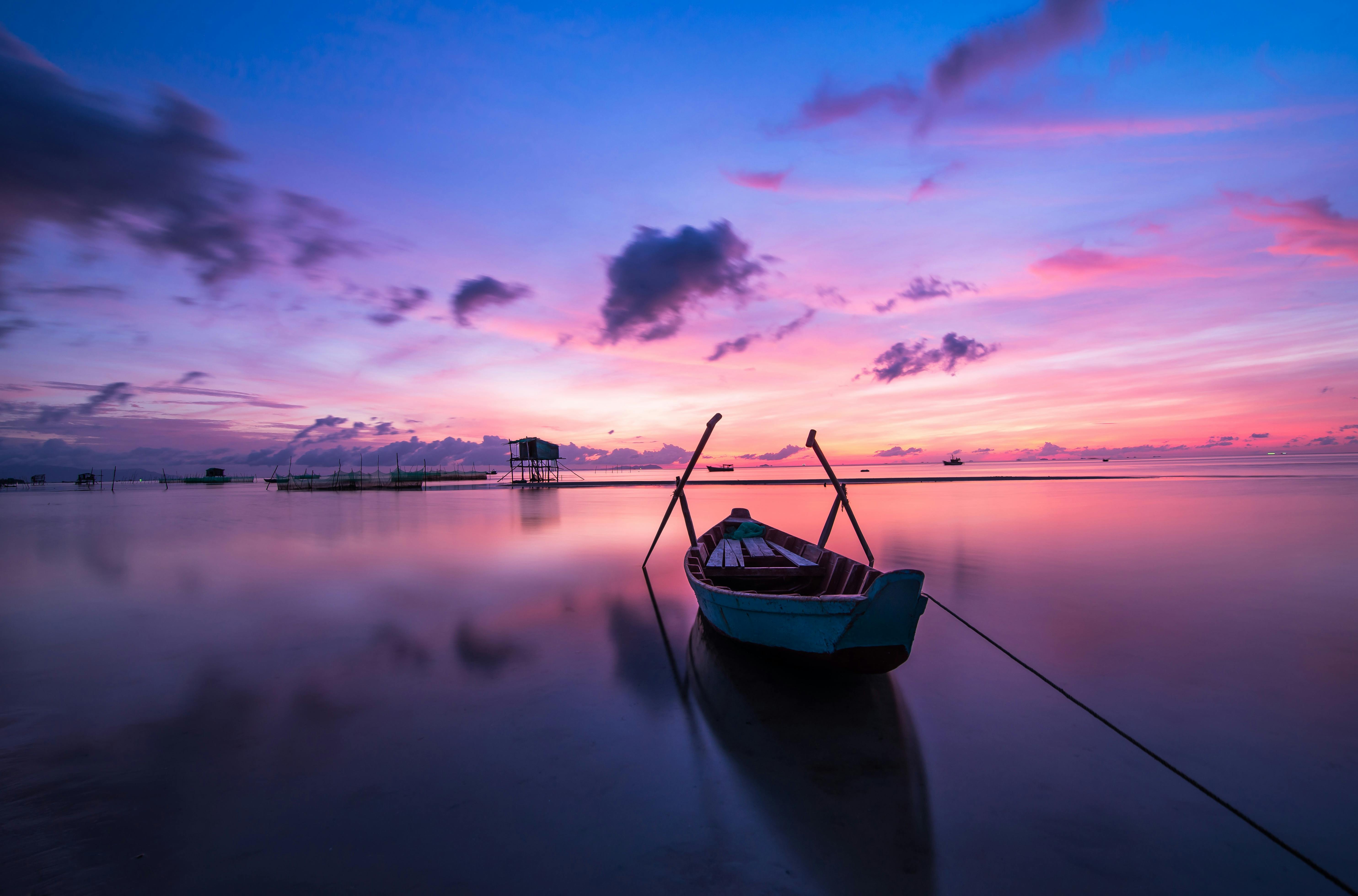 A small boat is anchored in the calm sea, the sky is dyed with beautiful pink and purple, with a few boats in the distance, the scene is peaceful and serene. - free wallpaper image