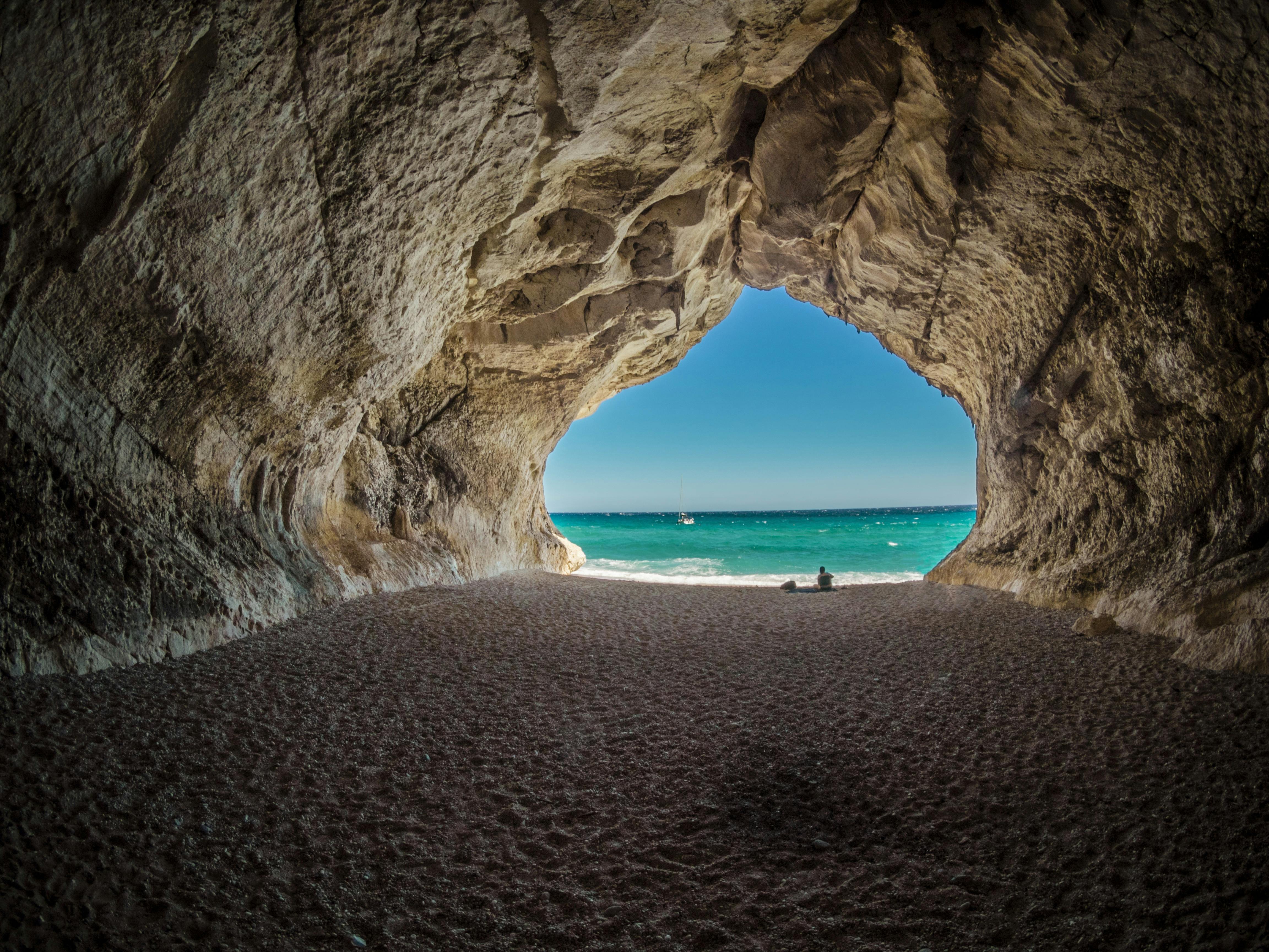 Inside a cave, you can see the beach and blue sky outside through the opening, a couple on the sand. - free wallpaper image