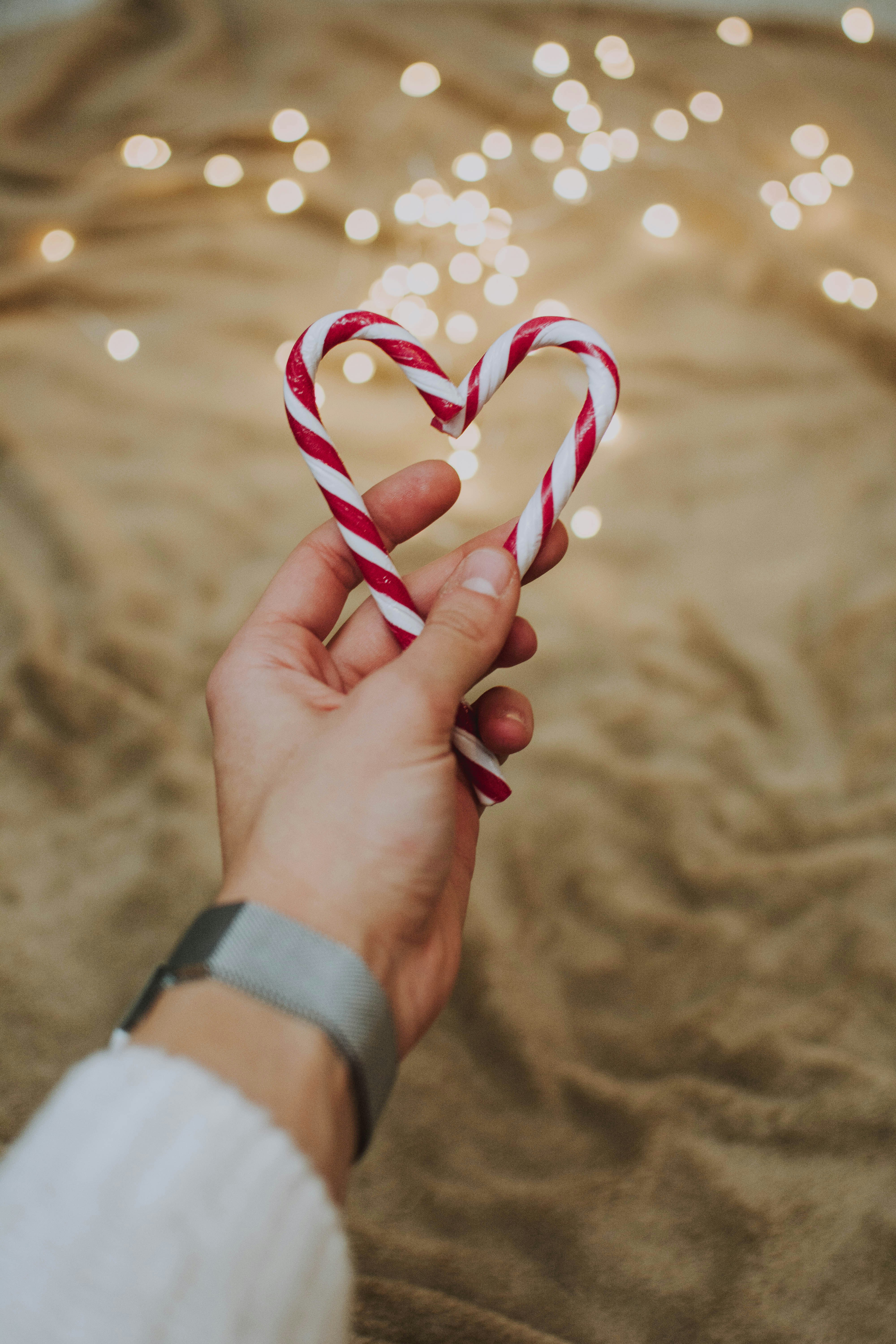 A hand holding two red candy canes, shaped into a heart, against a gold fabric background with small light bulbs. - wallpaper image