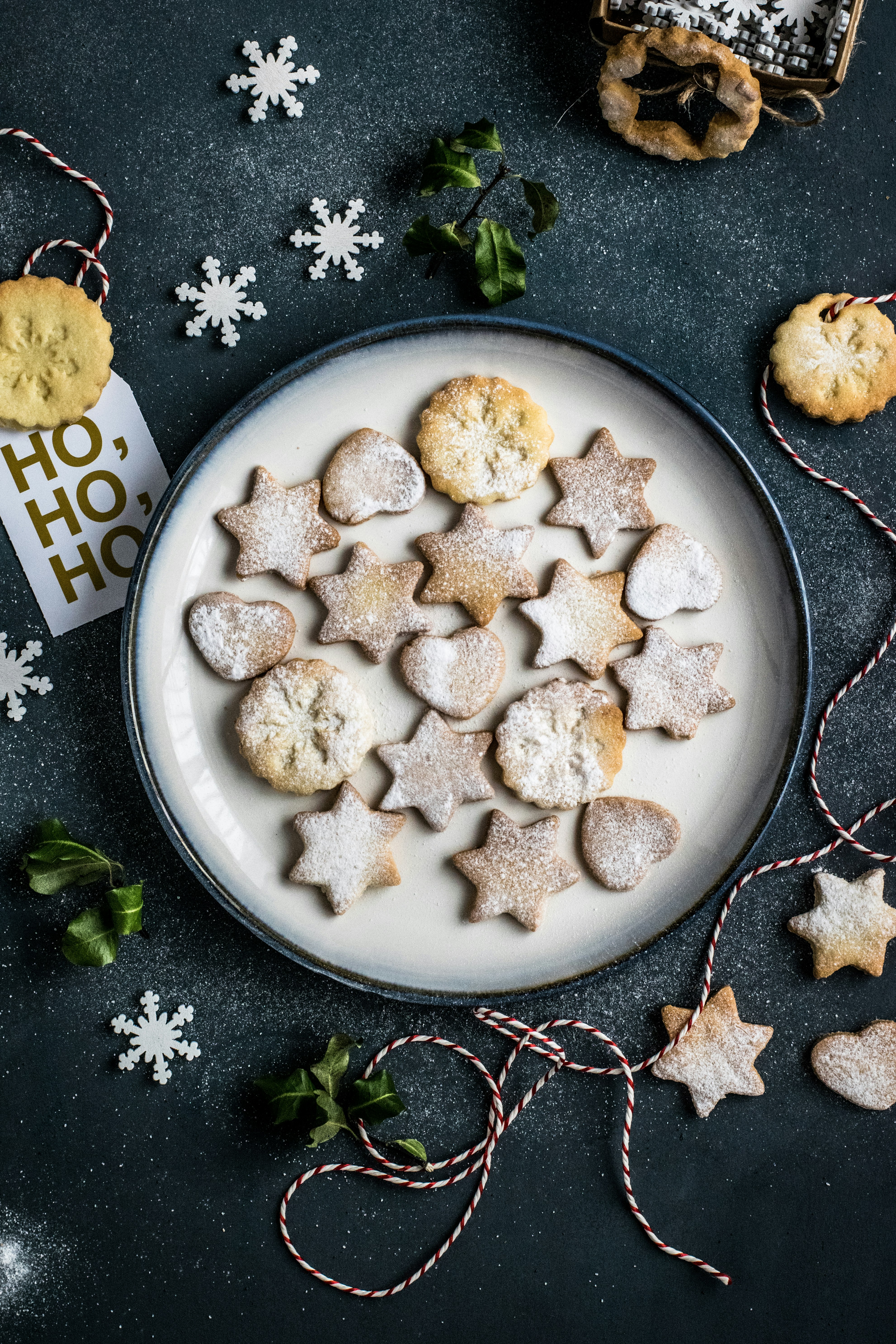 A white plate with many Christmas cookies decorated with powdered sugar, surrounded by red and white ropes, snowflake decorations and green leaves. - wallpaper image