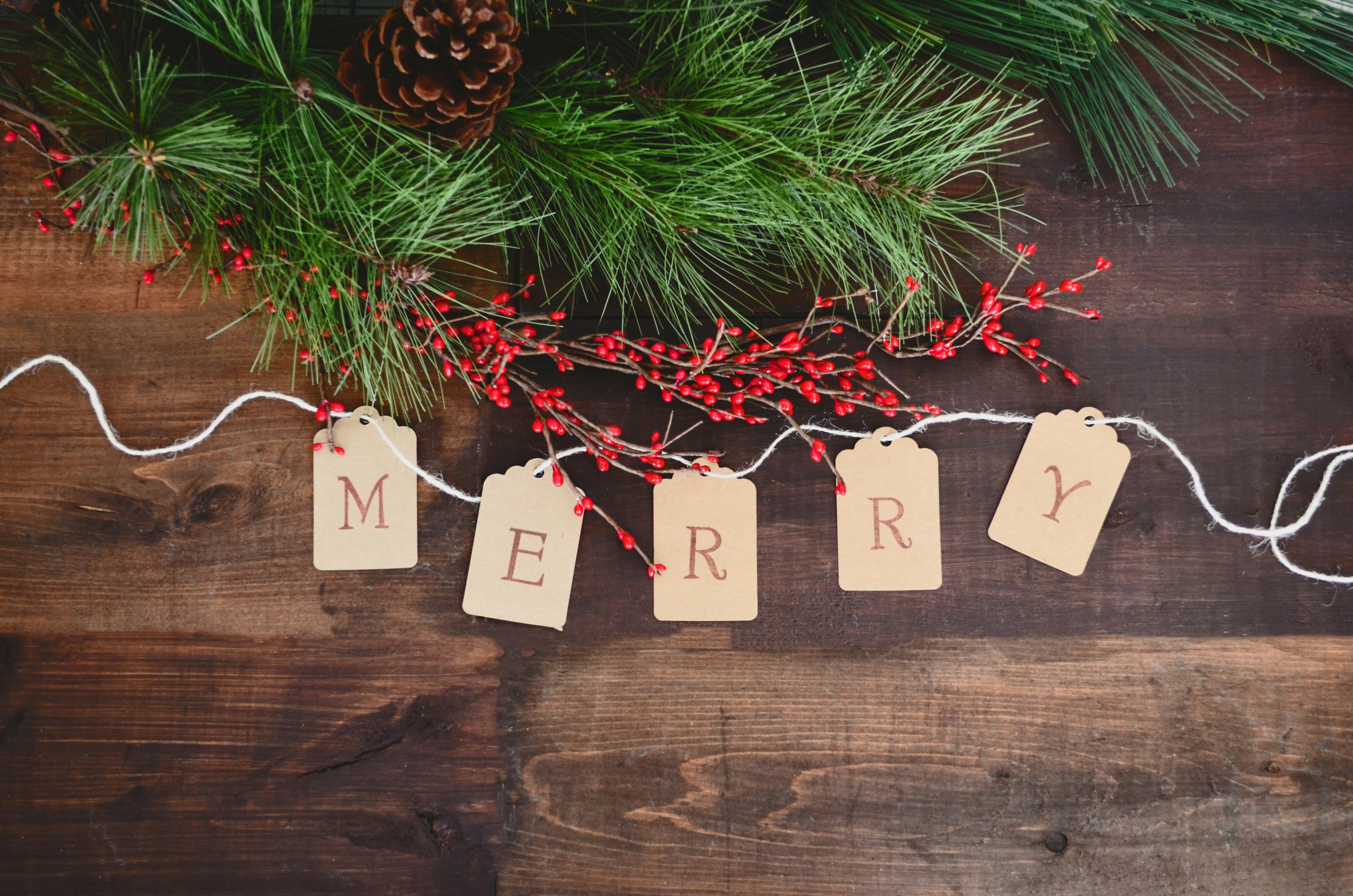 A brown wooden background decorated with green pine branches and red berries, with paper tags strung with twine that spell out "MERRY". - wallpaper image