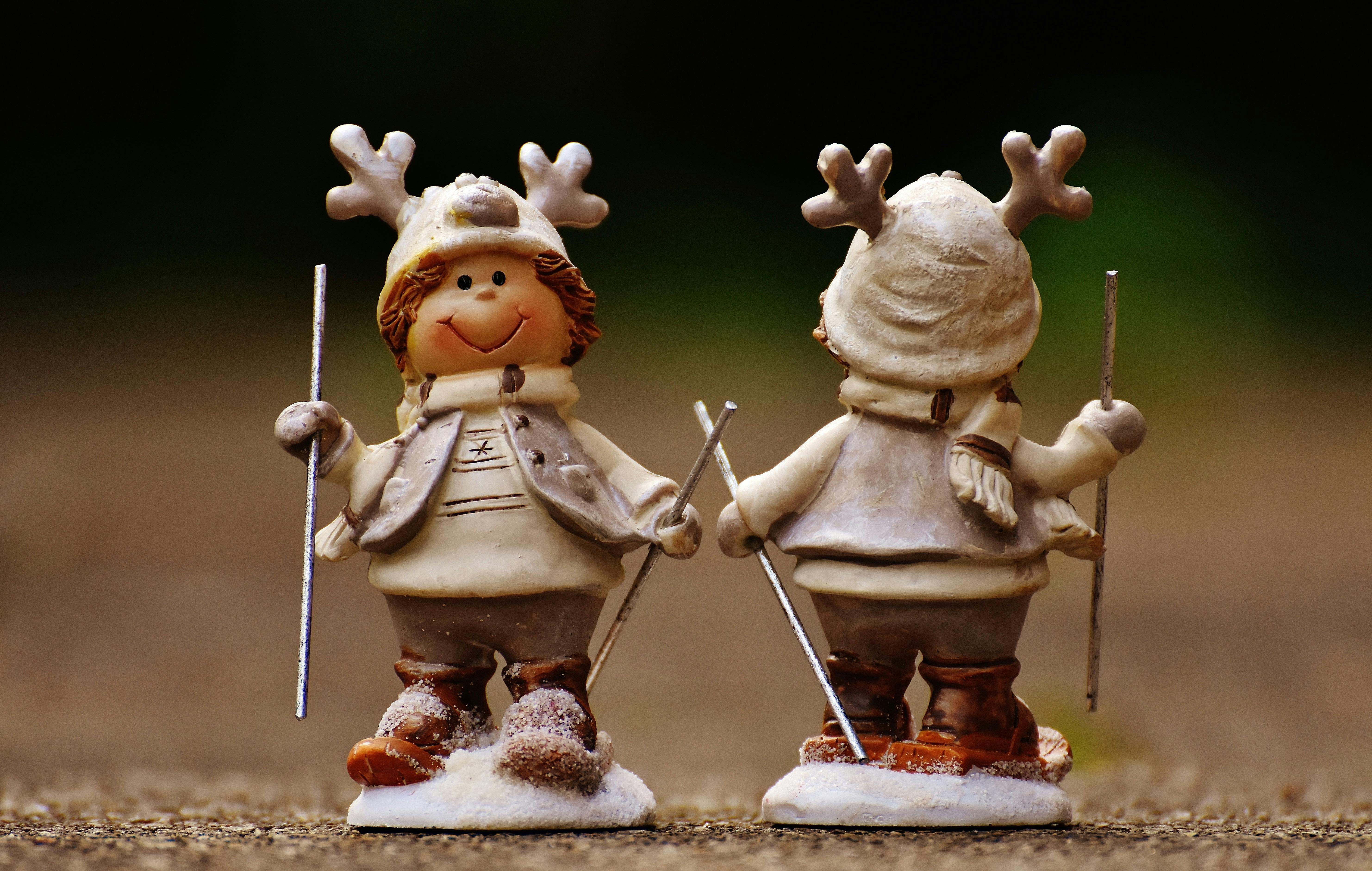 Two small figurines wearing white clothes and antlers, facing away from each other, holding ski poles and standing on snow. - wallpaper image