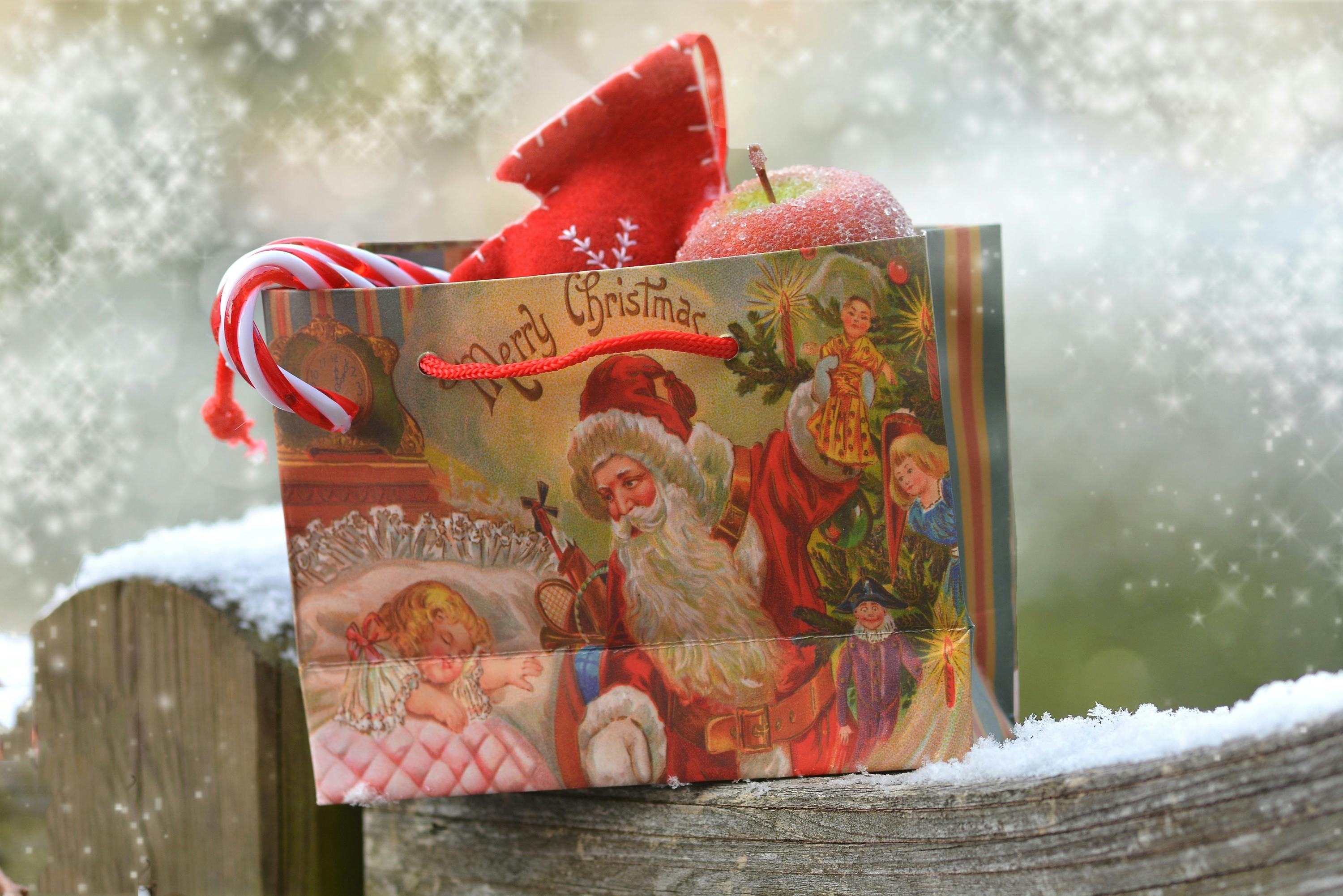 A red Christmas gift bag with Santa Claus and children printed on it, with a candy cane and an apple beside it, placed on the snow. - wallpaper image