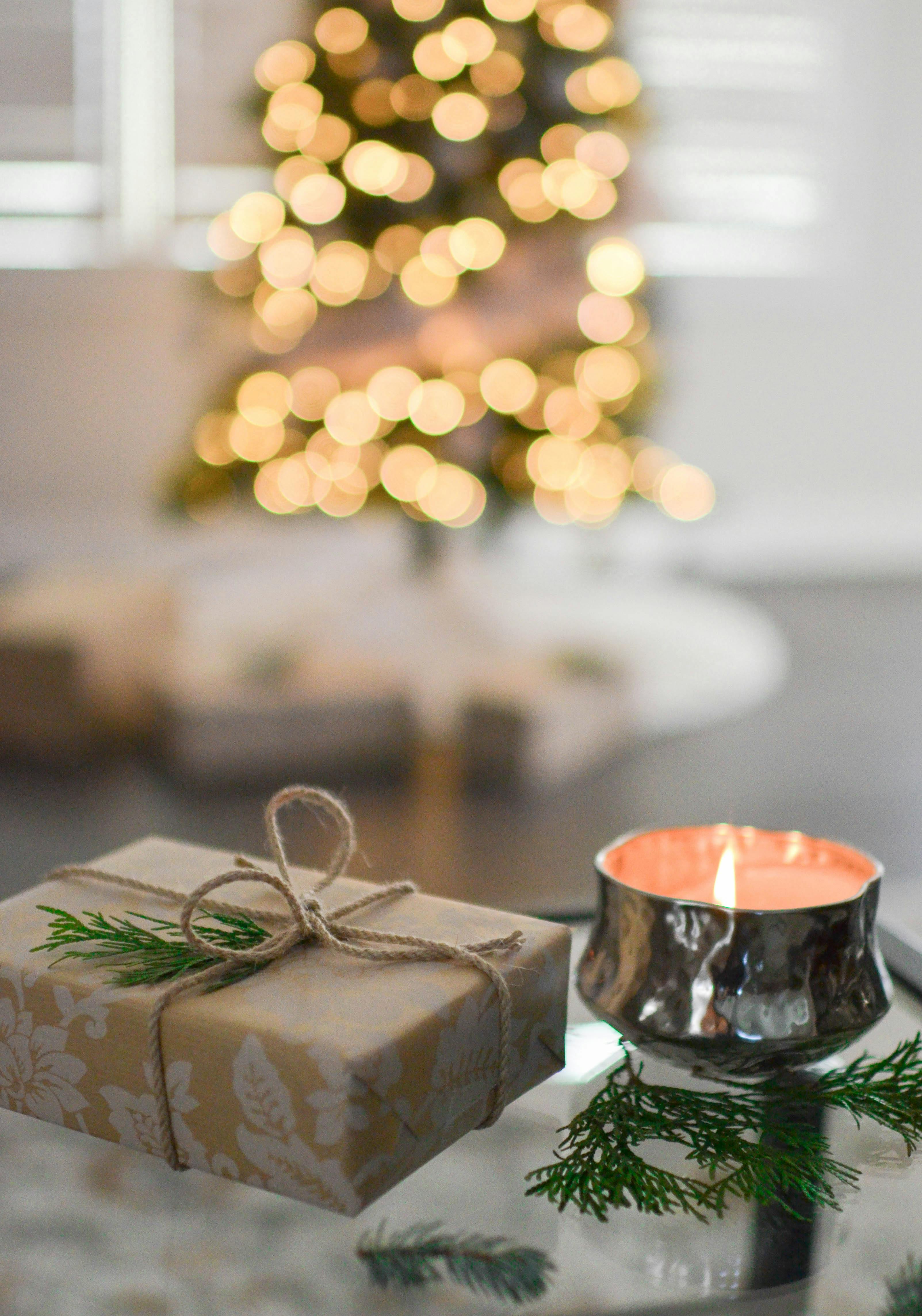 A beautifully wrapped gift box with a lit candle next to it, with a Christmas tree decorated with colorful lights in the background. - wallpaper image