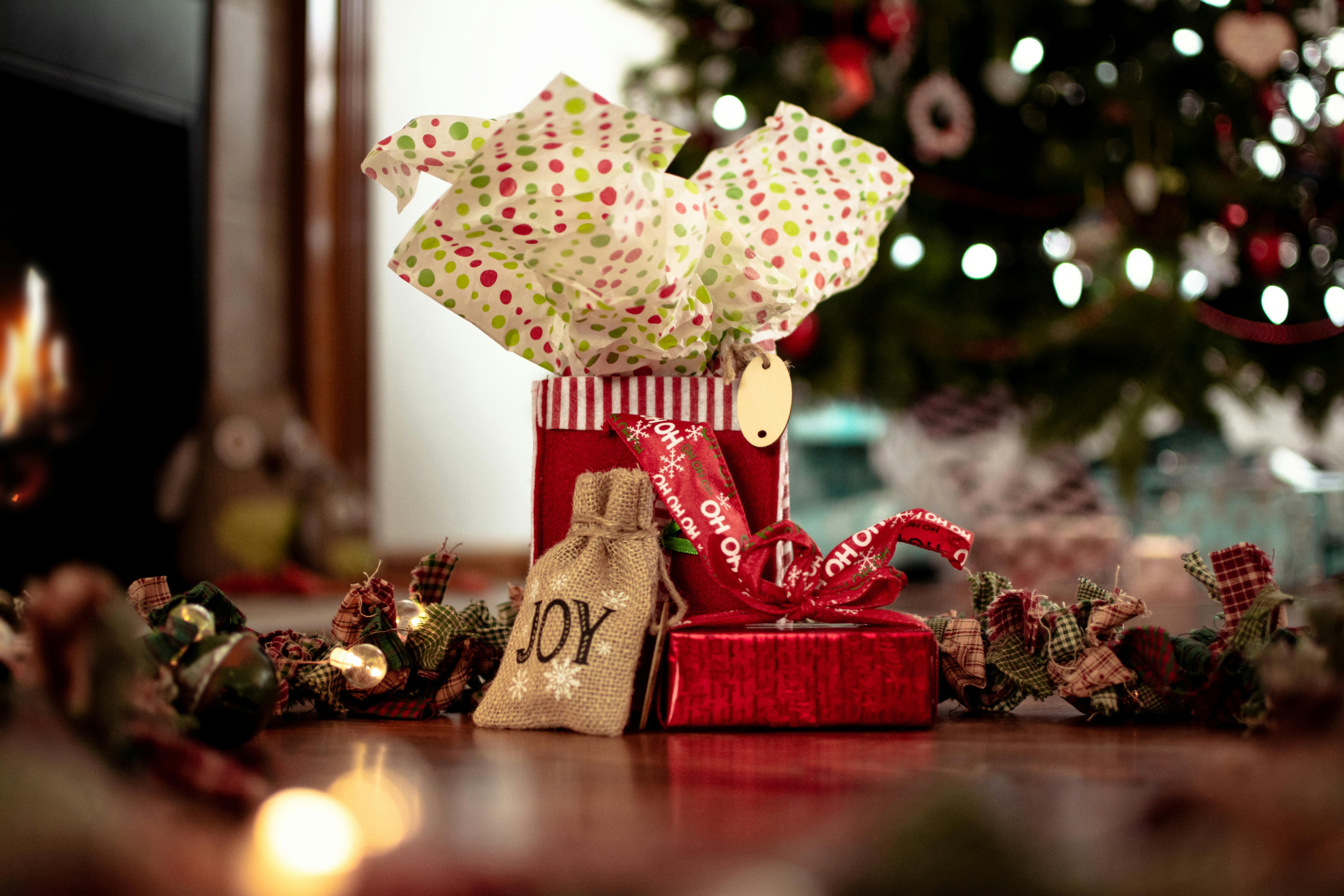 A Christmas gift box decorated with a red ribbon and a burlap bag with the word "joy" on it, with a decorated Christmas tree in the background. - wallpaper image