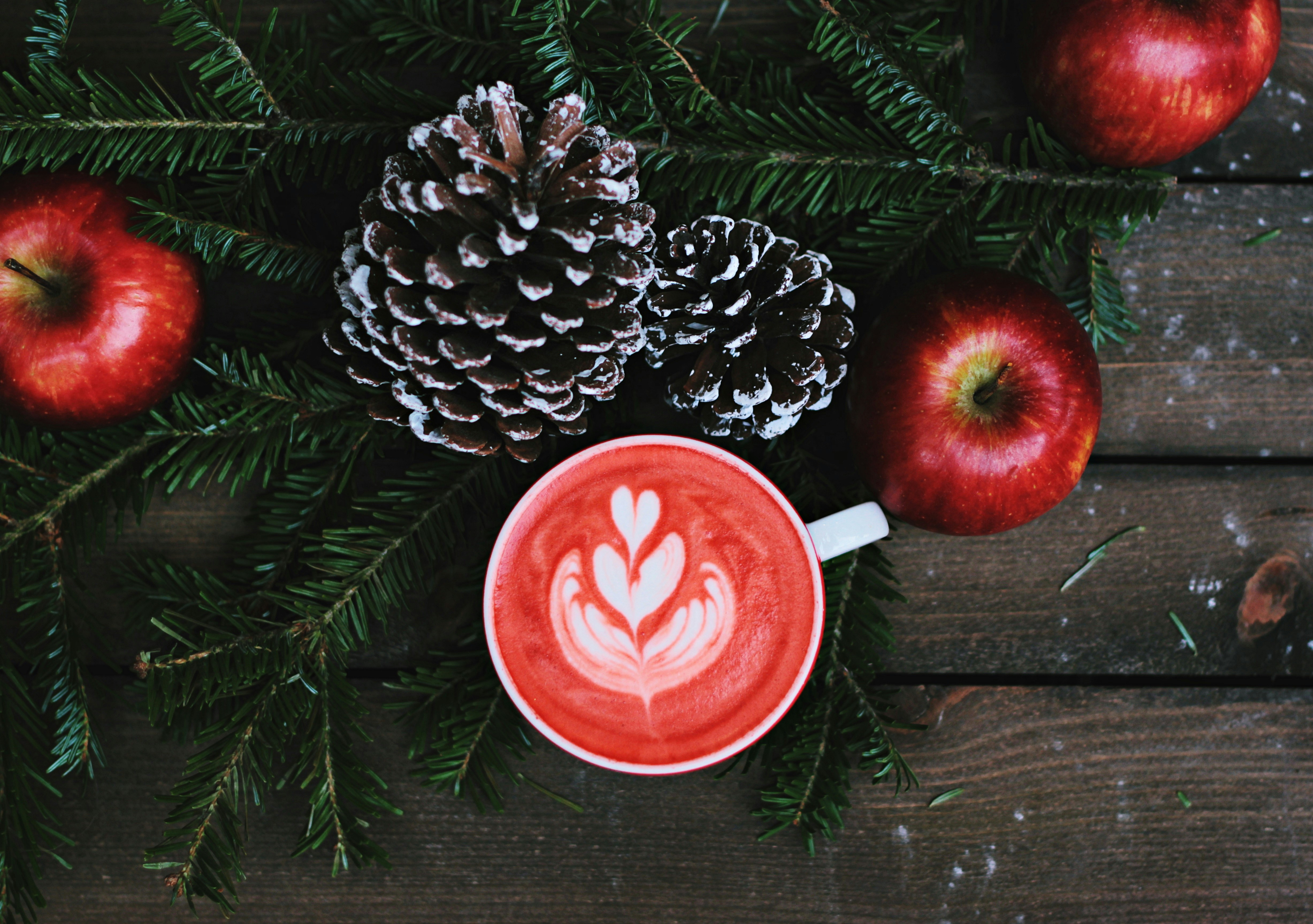 A red drink with white latte art, surrounded by pine branches, pine cones and red apples. The background is a wooden table. - wallpaper image