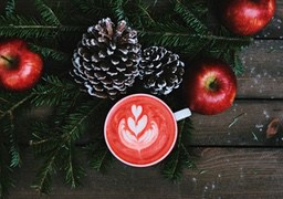 A red drink with white latte art, surrounded by pine branches, pine cones and red apples. The background is a wooden table. - wallpaper image