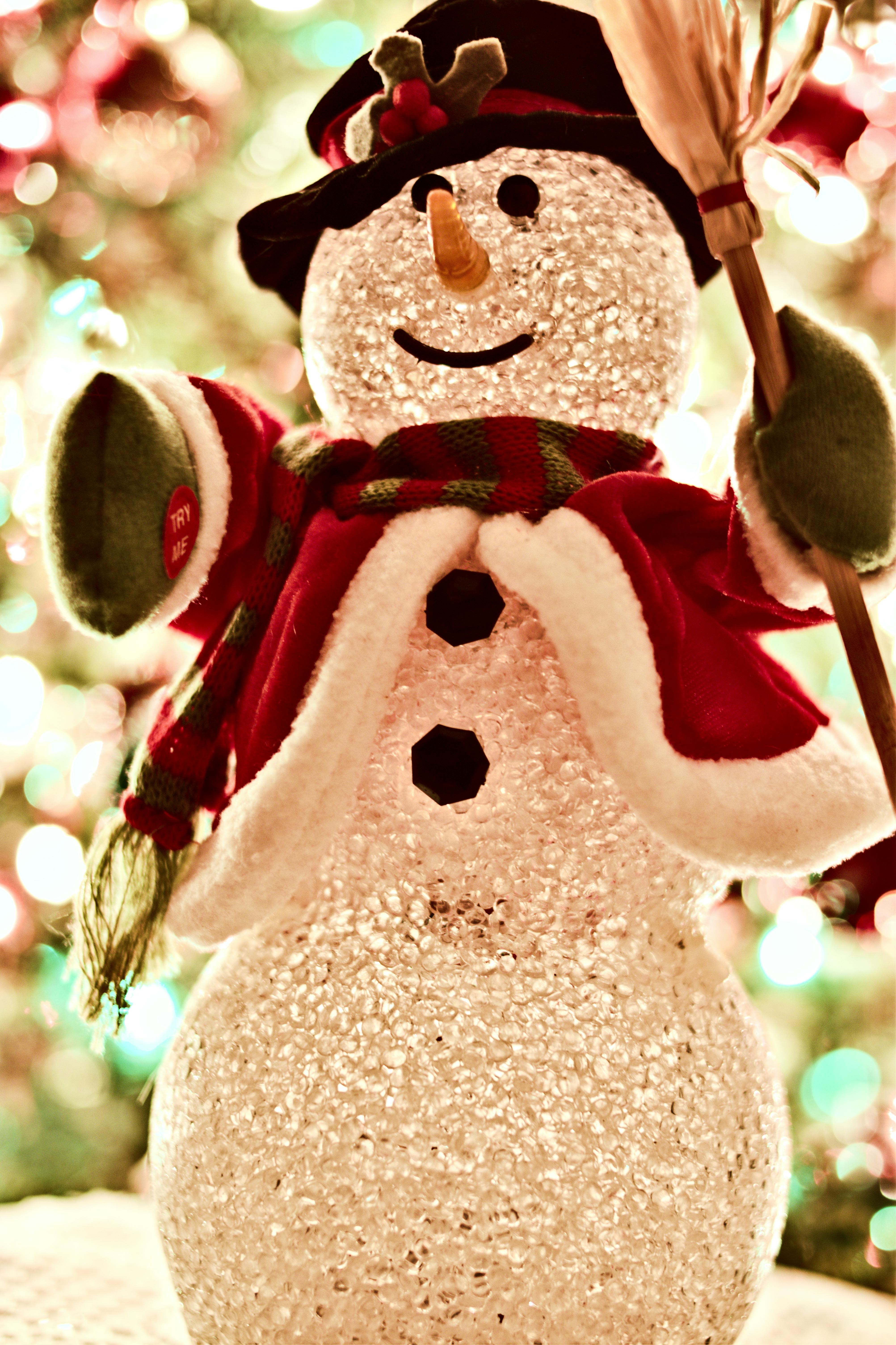 A snowman wearing a red coat and a black top hat stands in front of a Christmas tree, smiling at the camera. - wallpaper image