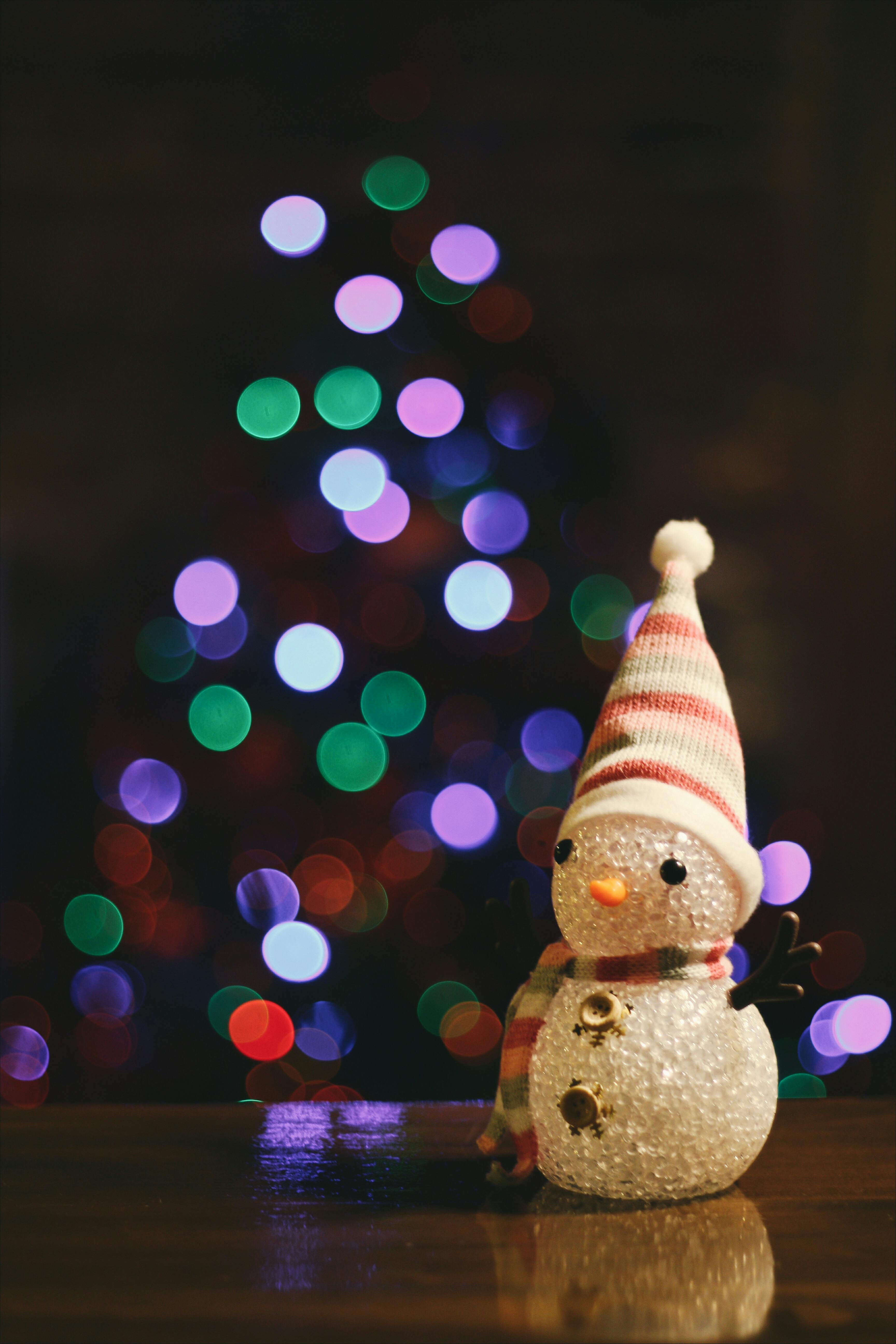 A snowman wearing a red and white striped hat stands in front of a Christmas tree with twinkling lights. The background is blurred, creating a warm festive atmosphere. - wallpaper image