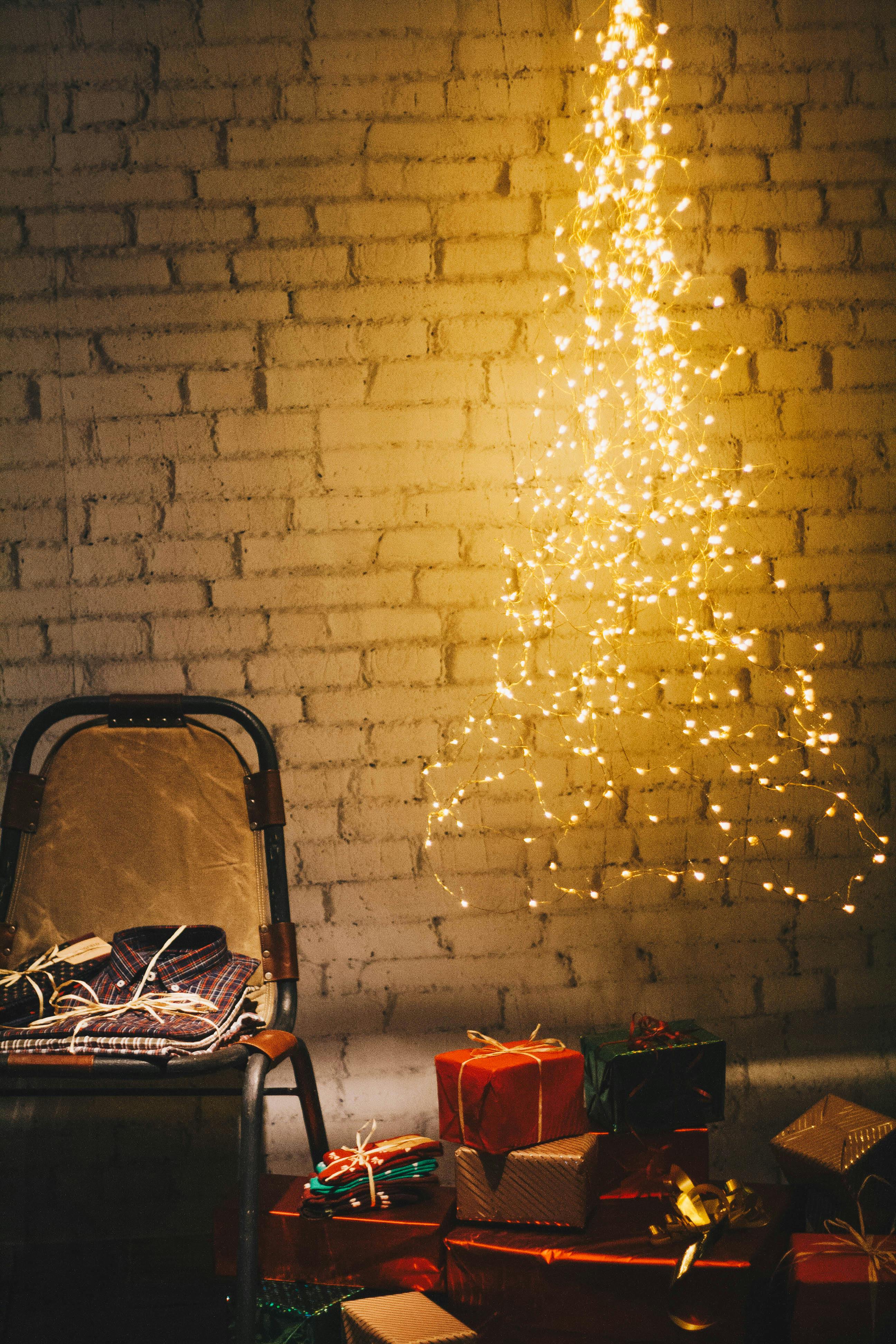 A wooden chair with some clothes on it, some gifts wrapped in colorful paper next to it, a Christmas tree decorated with fairy lights next to it, against a white brick wall. - wallpaper image