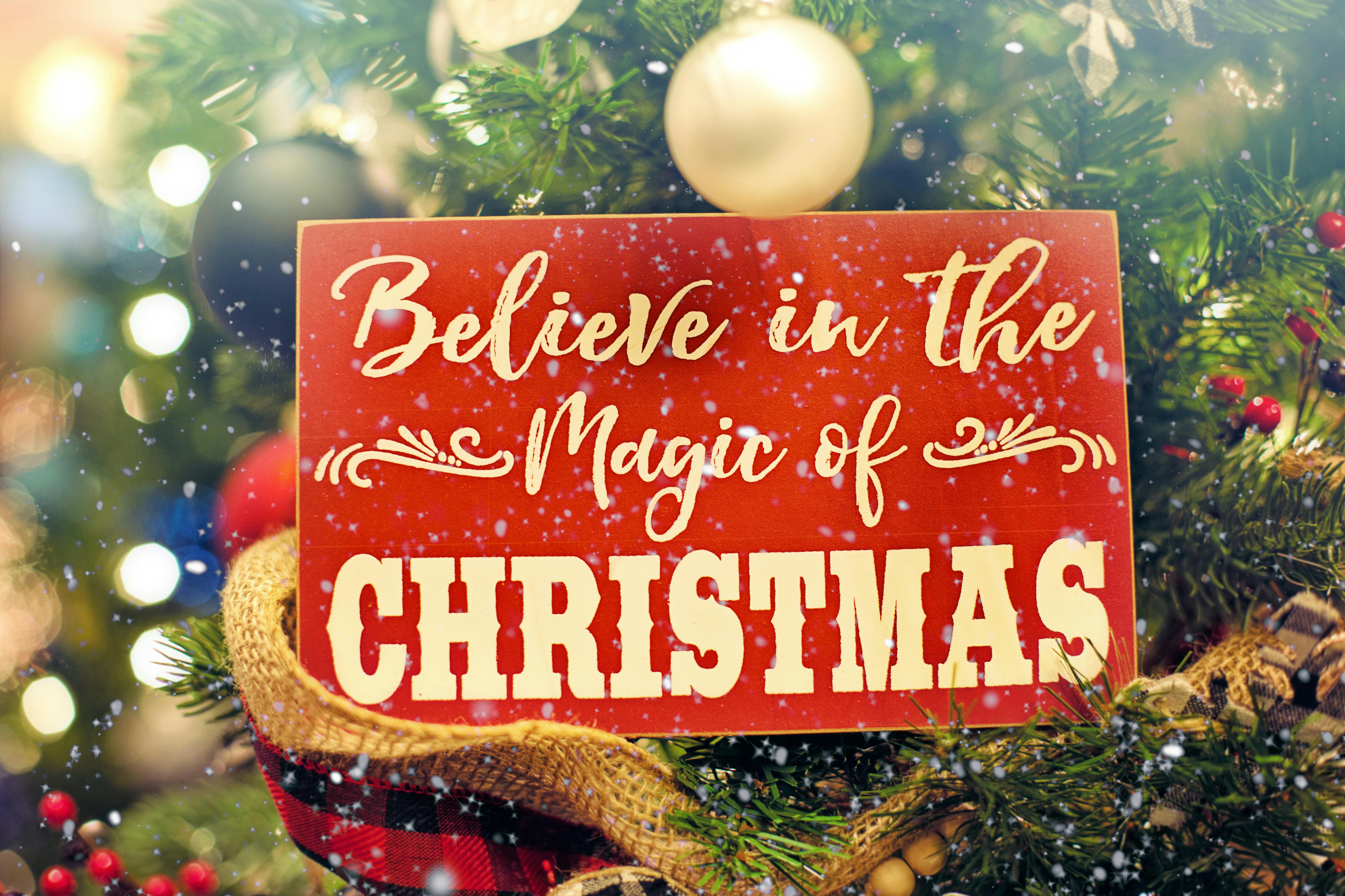 A red wooden sign that says "Believe in the Magic of Christmas" hangs on a Christmas tree decorated with golden ornaments and green branches, creating a festive atmosphere. - wallpaper image