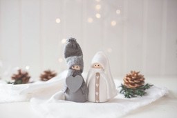 Two wooden dolls wearing sweaters stand on a white cloth, next to pine cones and green plants. - wallpaper image