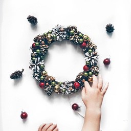 A child&apos;s hand is decorating a Christmas wreath with pine cones, green balls, red balls and golden decorations - wallpaper image