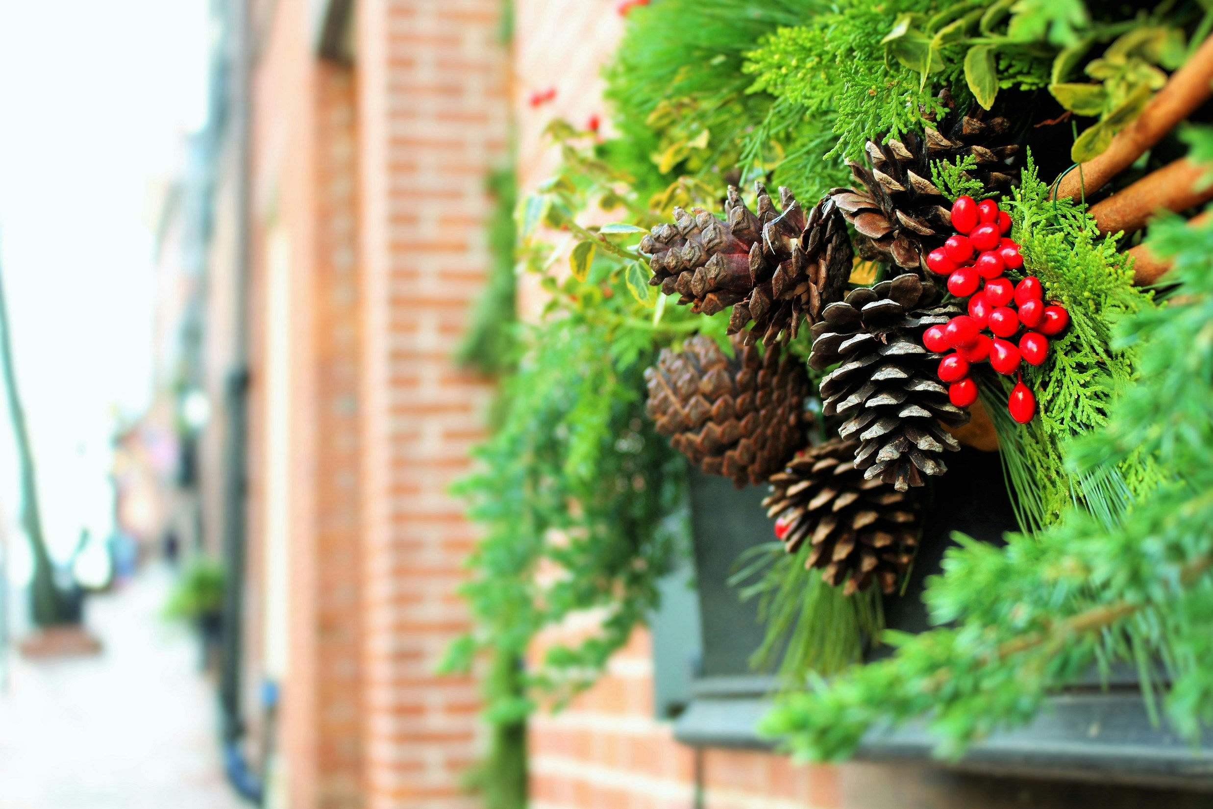 A Christmas wreath decorated with pine cones, red berries and green leaves, against a blurry red brick wall background. - wallpaper image