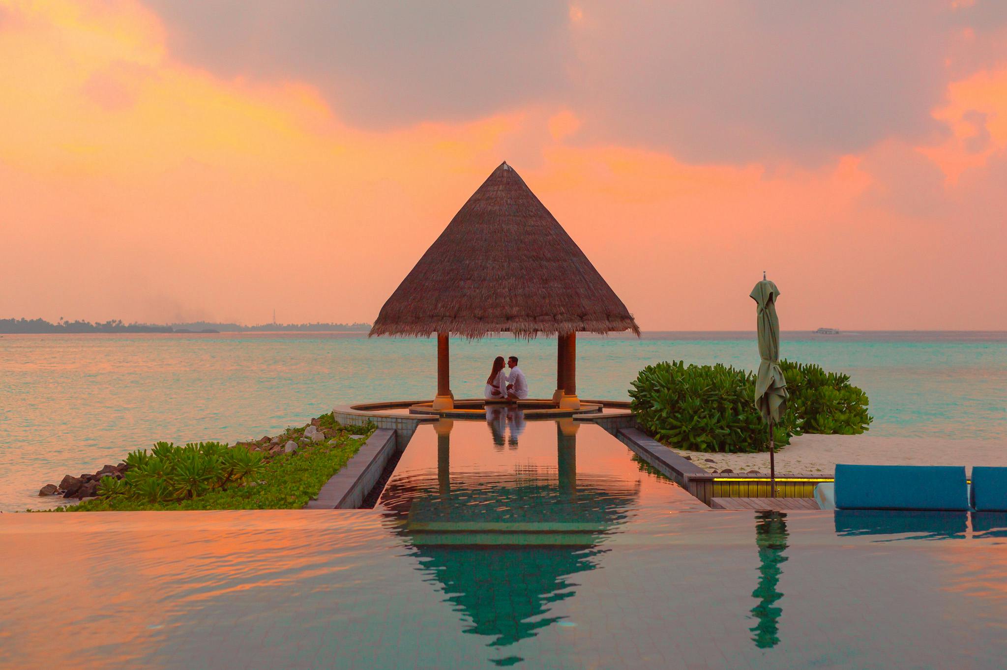 A couple relaxing by the pool at a resort on the beach, the sky is a beautiful orange and pink as the sun sets, the water is clear and blue, the scenery is picturesque. - free wallpaper image