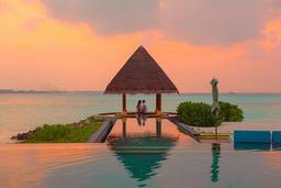 A couple relaxing by the pool at a resort on the beach, the sky is a beautiful orange and pink as the sun sets, the water is clear and blue, the scenery is picturesque. - free wallpaper image