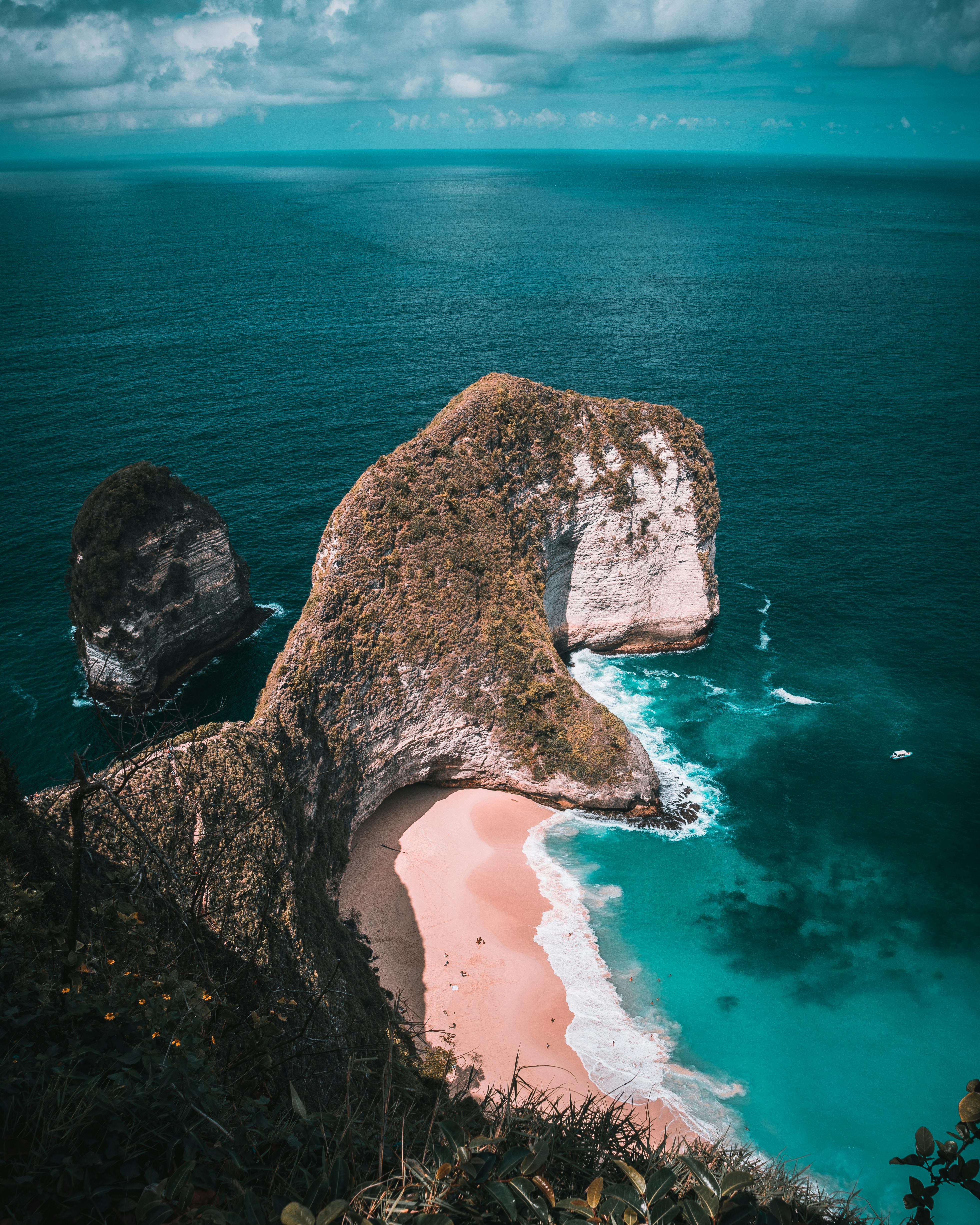 An overhead view of a small cove by the sea, with a sandy beach and some tourists, the cove is surrounded by towering rocks with green plants, and the vast blue sea extends outwards. - free wallpaper image