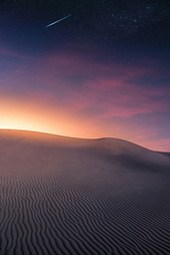 A shooting star appears in the sky over a white sand dune in the desert, against a background of purple, pink and orange night sky with stars. - free wallpaper image