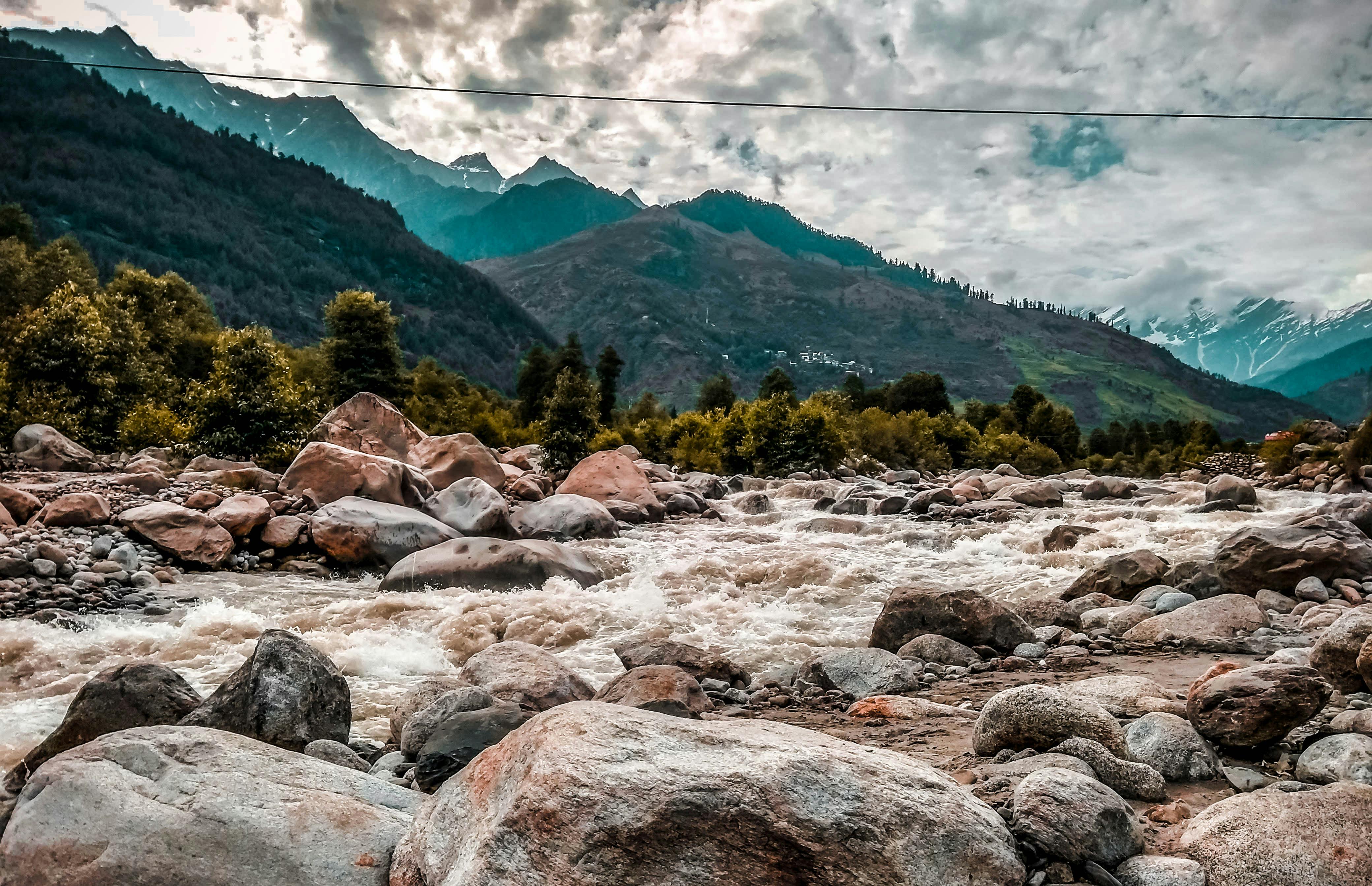 A fast-flowing river in the mountains, the riverbed is covered with rocks of different sizes, and in the distance are rolling green hills. - free wallpaper image