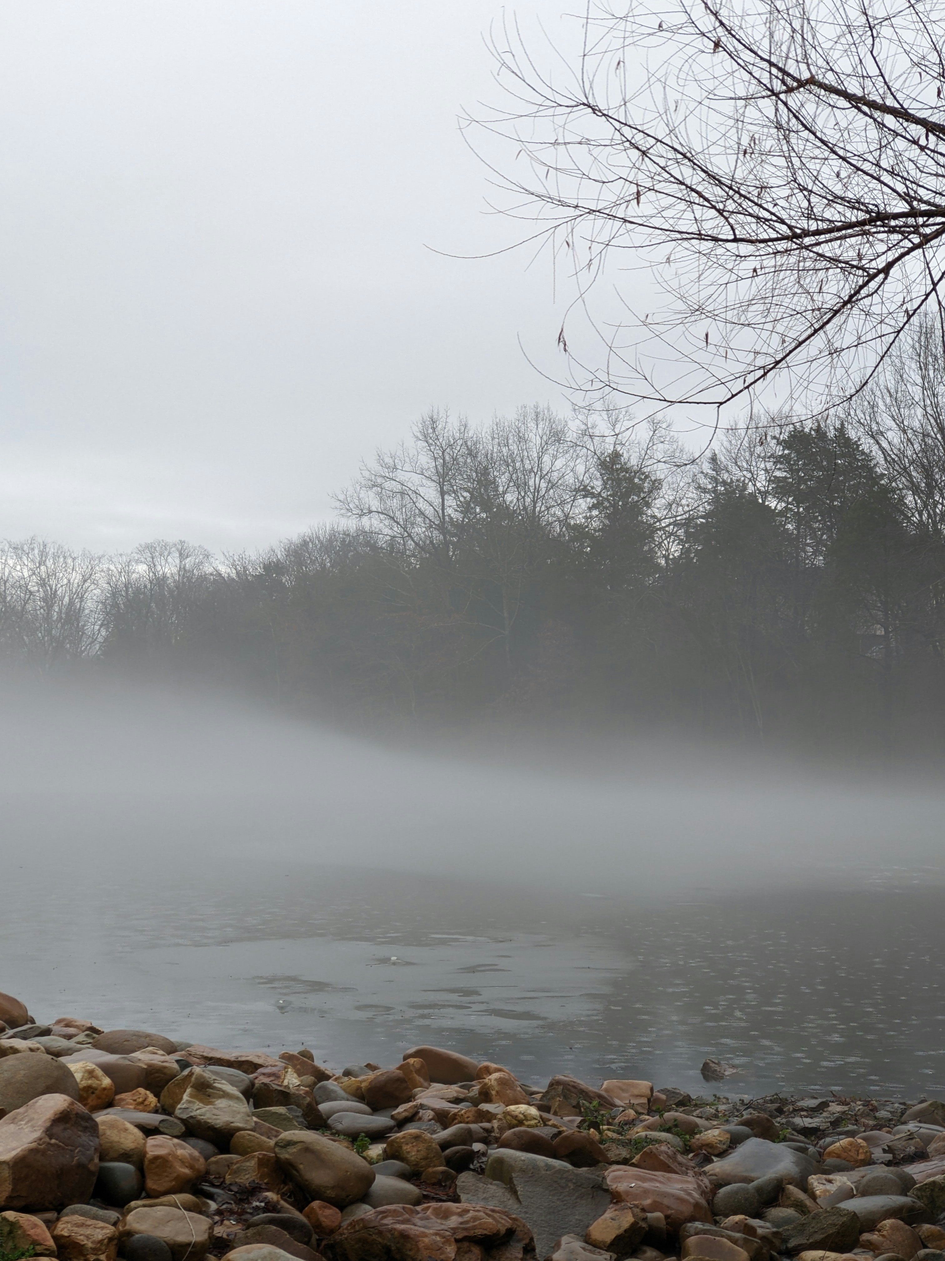 A river is covered in fog, with calm water and a shoreline of pebbles of various sizes. - free wallpaper image