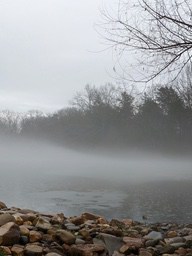 A river is covered in fog, with calm water and a shoreline of pebbles of various sizes. - free wallpaper image