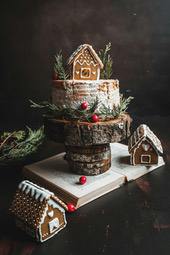 A gingerbread cake decorated with Christmas tree branches and berries, placed on a wooden round plate, the wooden round plate is placed on an open book, and there are three gingerbread houses around it. - wallpaper image