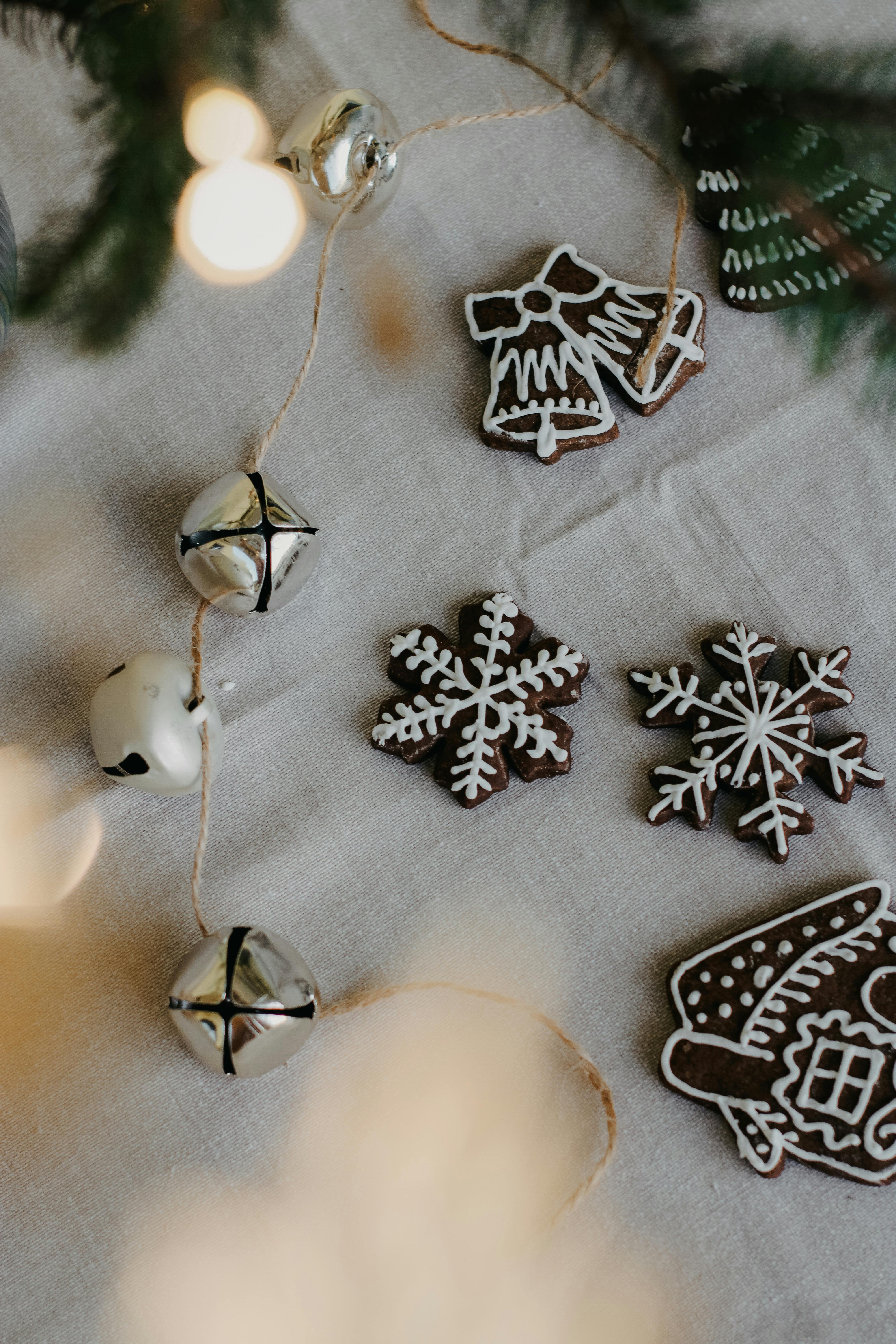 There are some gingerbread cookies on a white tablecloth, the shapes of the cookies are Christmas trees, bells, snowflakes and houses, and there are silver bells decorations beside them. - wallpaper image