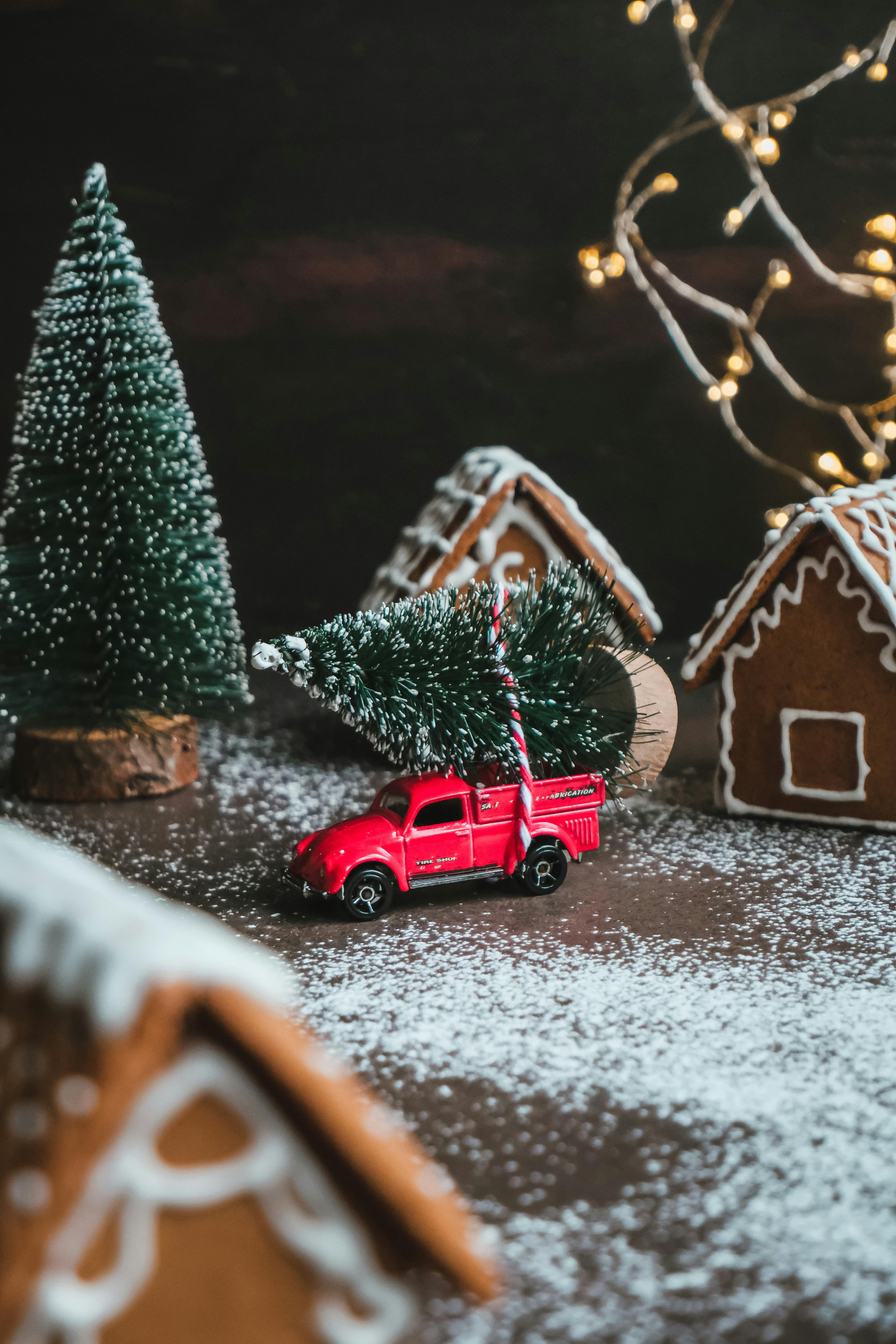 A red toy car carrying a small Christmas tree, next to gingerbread houses, the ground is covered with powdered sugar, creating a winter snow scene atmosphere. - wallpaper image