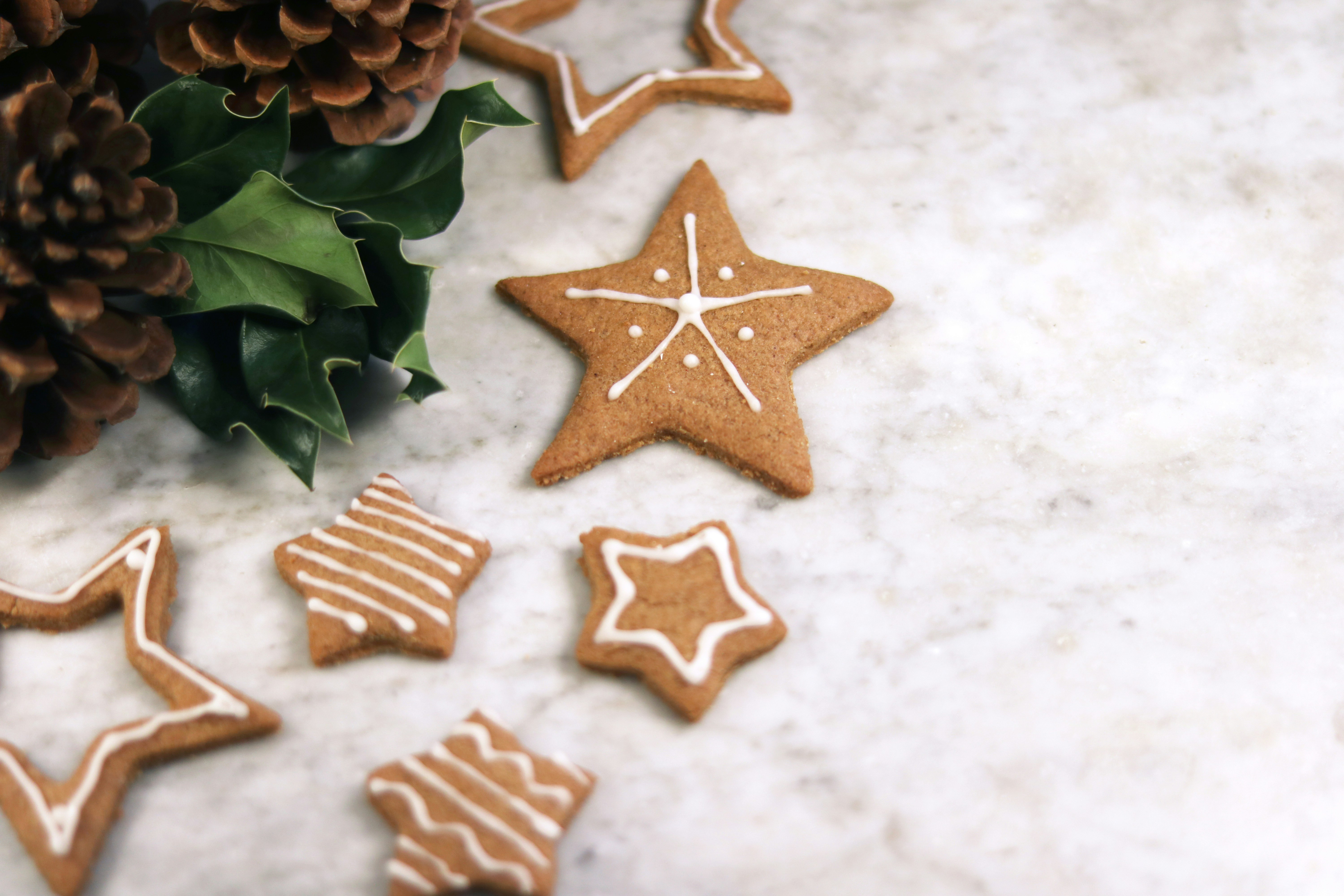 Several gingerbread stars are scattered on a white marble table top, with pine cones and holly leaves beside them. - wallpaper image