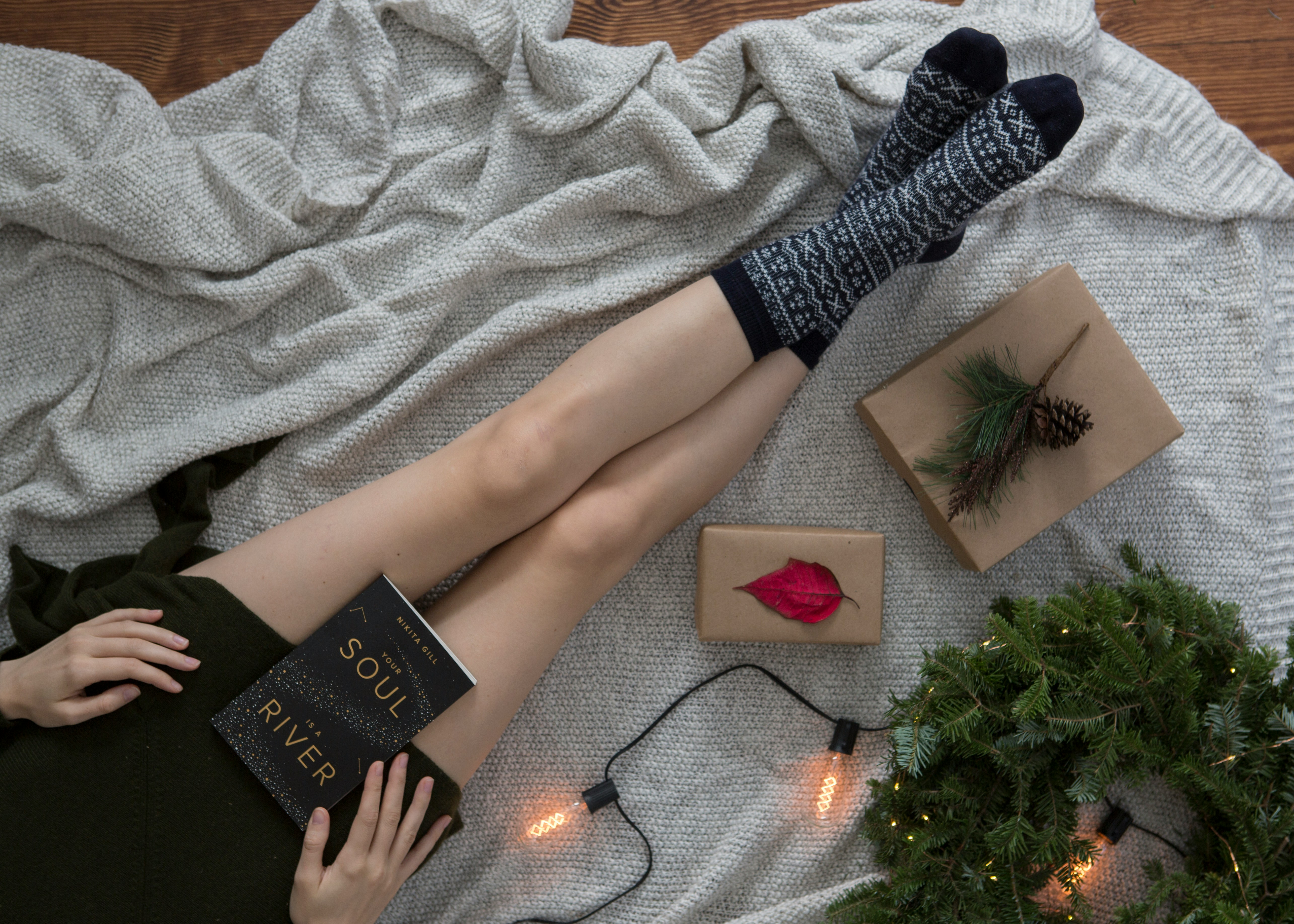 A girl in a green sweater lies on a white blanket with a book in her hands, wearing black and white patterned socks, next to a gift box, a Christmas tree and some decorative lights - wallpaper image