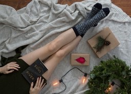 A girl in a green sweater lies on a white blanket with a book in her hands, wearing black and white patterned socks, next to a gift box, a Christmas tree and some decorative lights - wallpaper image