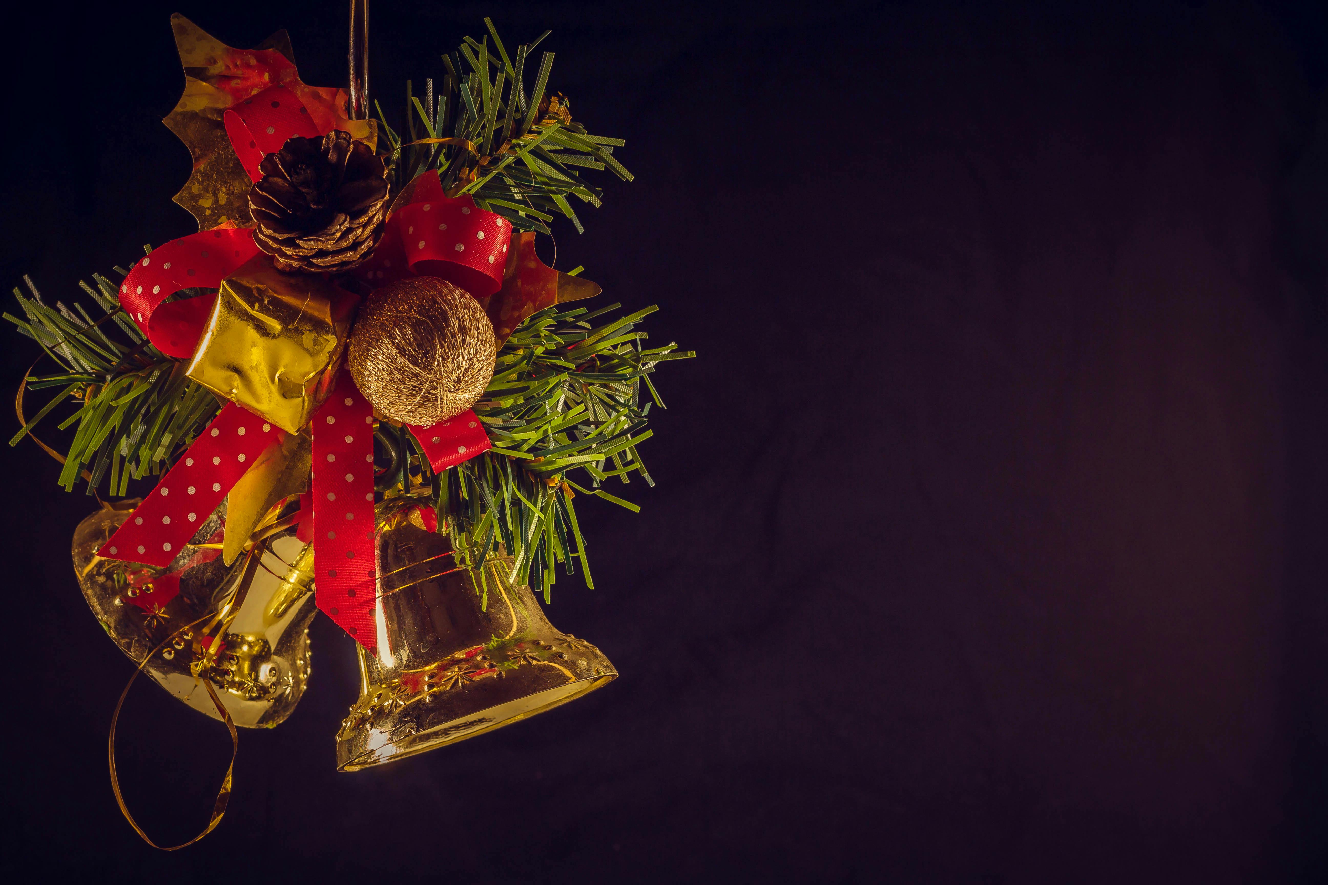 Two golden bells decorated with red ribbon and green pine branches hanging against a black background. - wallpaper image